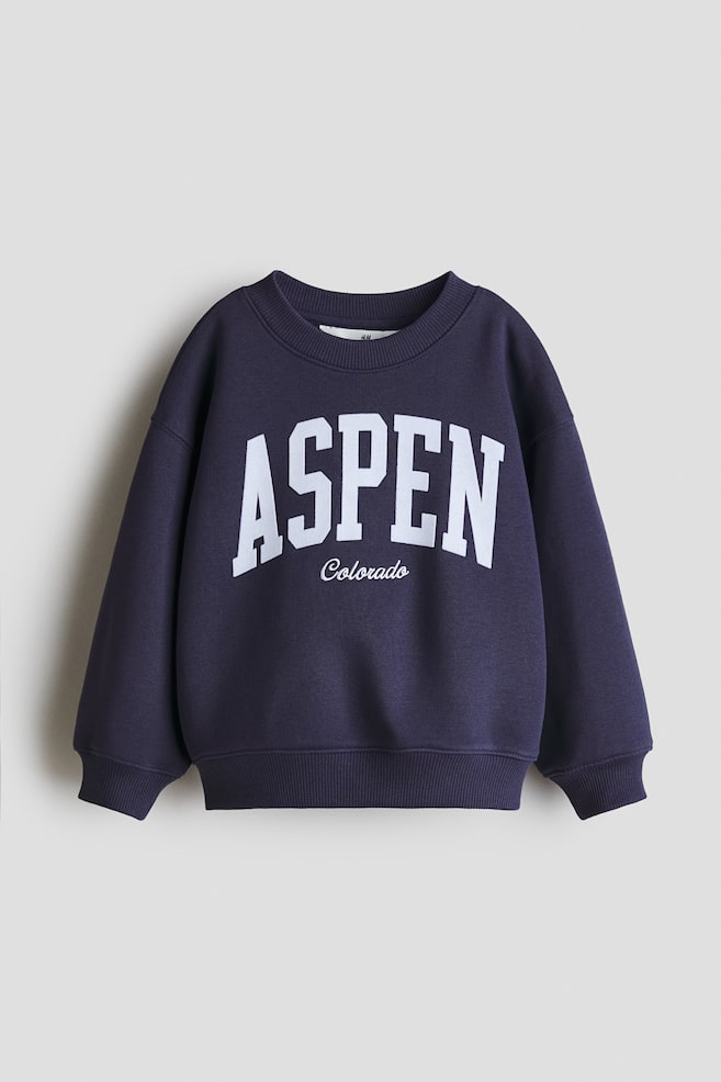 Un sweat-shirt bleu foncé présente son DEVANT, arborant une grande impression blanche "ASPEN" avec "Colorado" en script plus petit en-dessous. Il est conçu avec un col rond côtelé, des épaules tombantes, des poignets côtelés et un ourlet côtelé. - 1