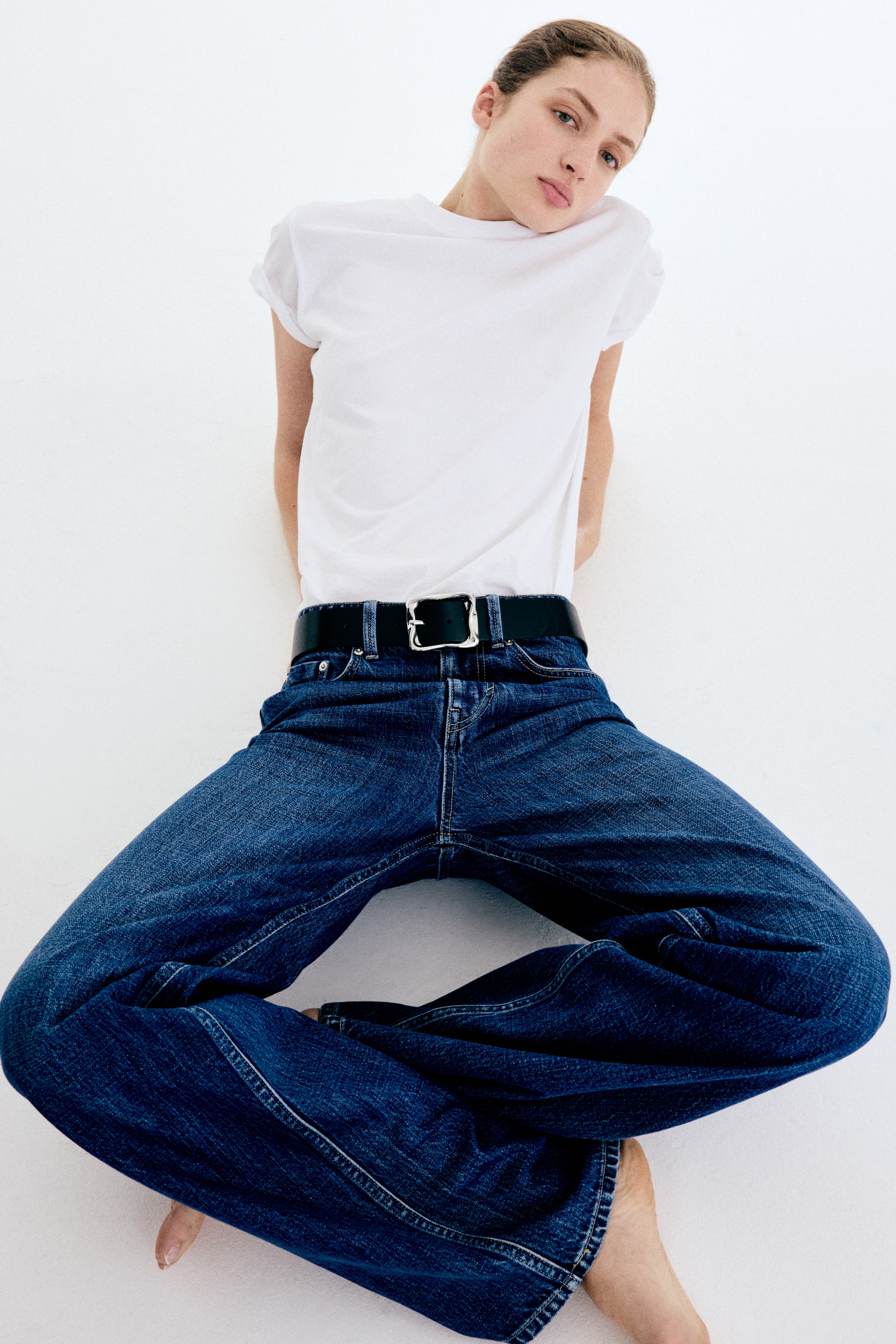 Se større billede: En hvid crew neck T-shirt med opsmøgede korte ærmer er indstoppet i mørkeblå wide-leg jeans. Jeansene er strammet ind i taljen af et sort bælte med et sølvfarvet, rektangulært spænde.