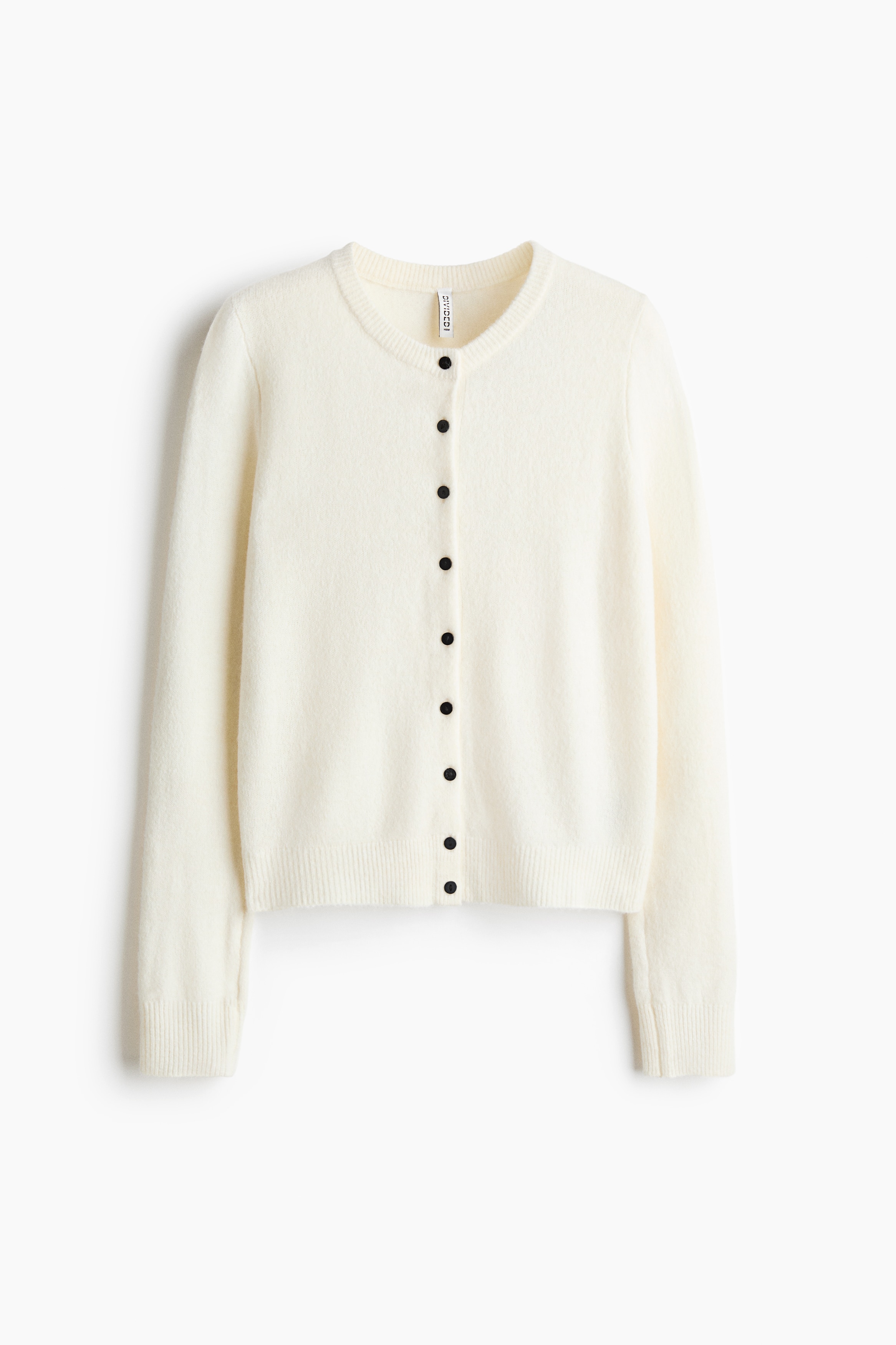 A cream-colored, soft-knit cardigan is being worn by the model, who is raising one long sleeve toward the foreground. The fitted cardigan features dark buttons down the front and a ribbed hem, and is styled with casual brown trousers.