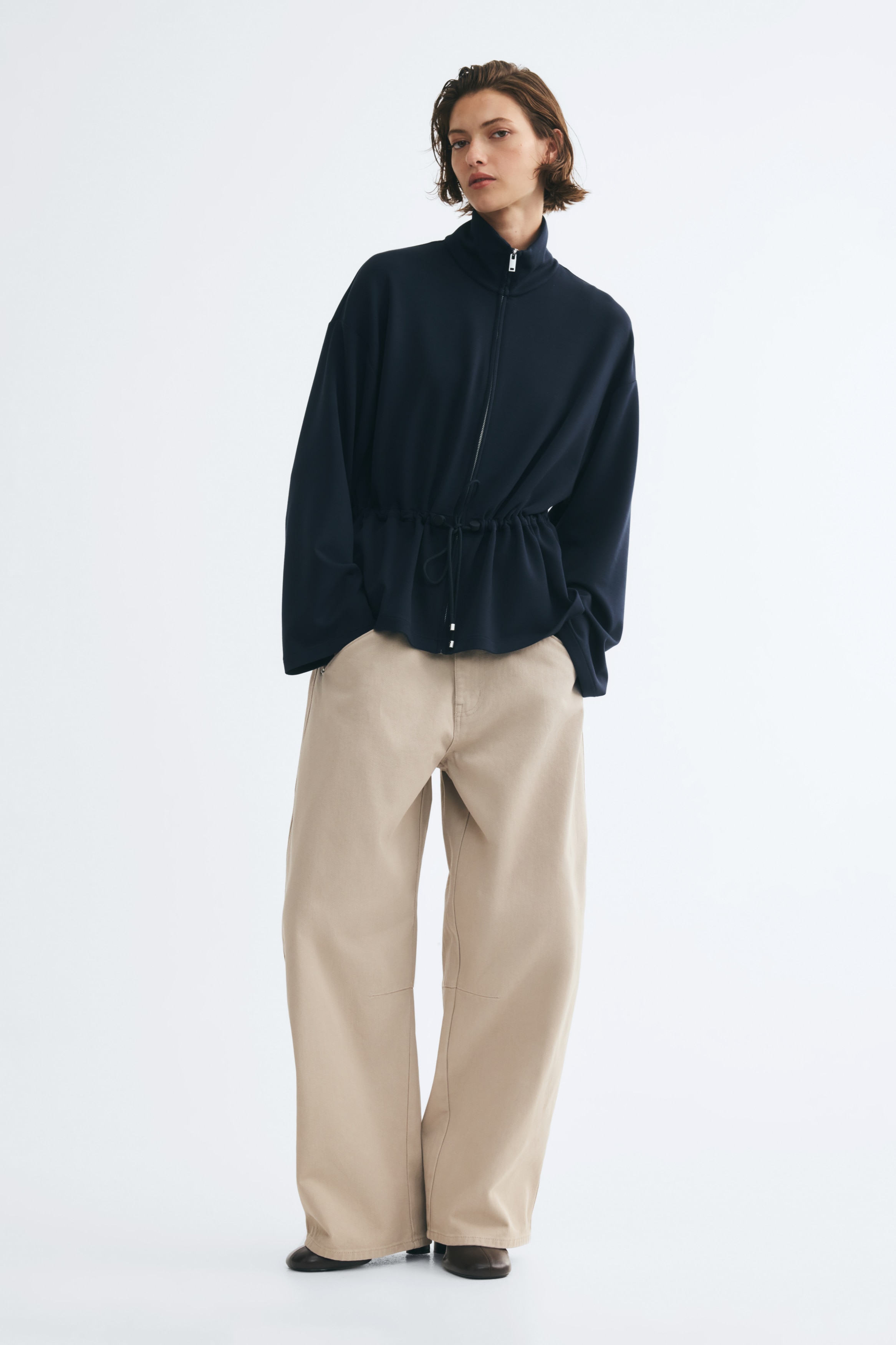 A dark blue zip-up jacket is facing front, featuring a stand-up collar and a full-length zip. It is displaying long, wide sleeves and a drawstring waist that is cinching the fabric and ending with silver-toned tips.