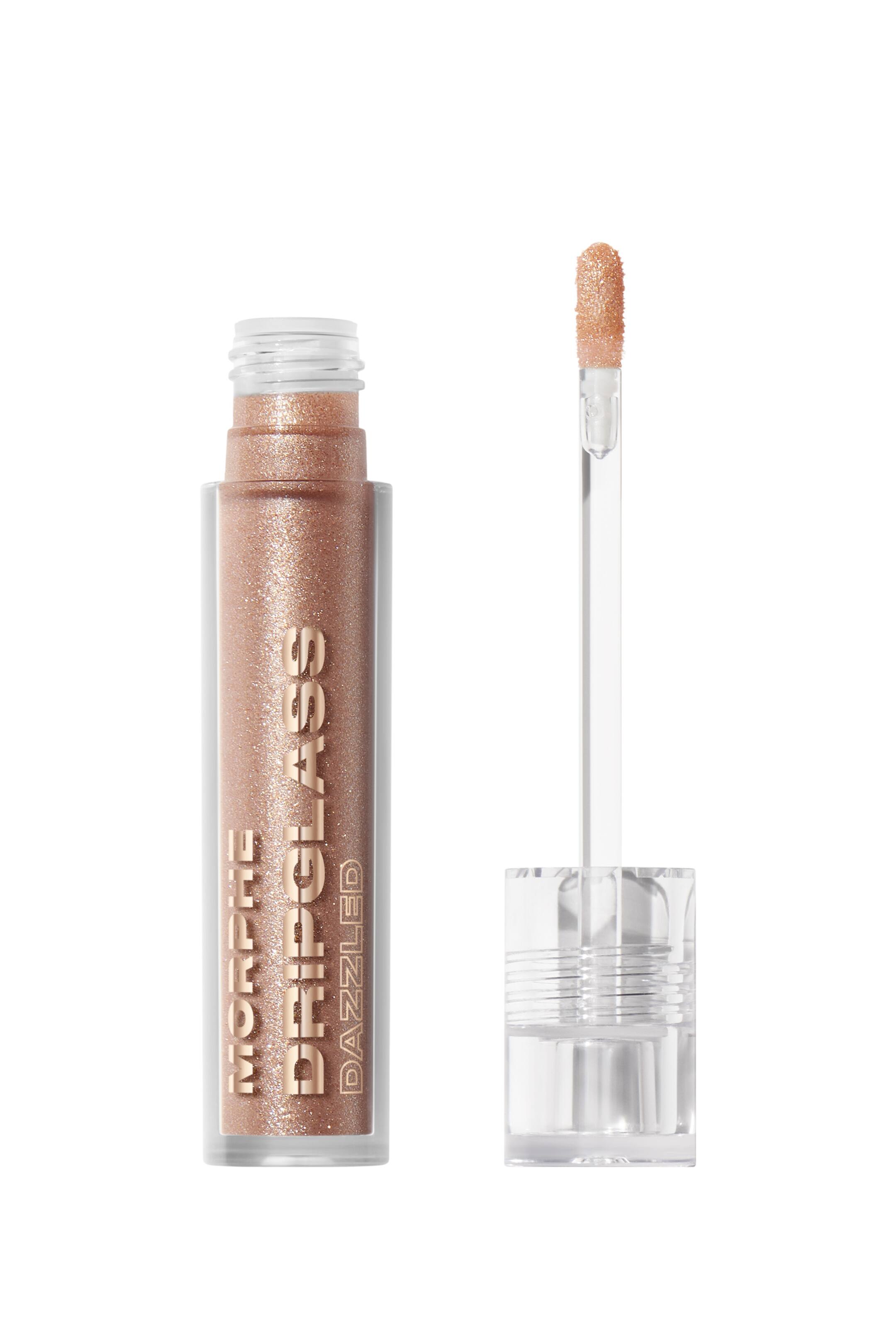 Visa större bild: En Morphe Dripglass Dazzled läppglans presenteras. Dess klara rektangulära tub är fylld med en skimrande roséguldfärgad produkt och visar "MORPHE" staplat ovanför "DRIPGLASS" vertikalt, med "DAZZLED" tryckt horisontellt nedanför på framsidan. En klar applikatorstav, med sin flockade spets täckt av samma skimrande produkt, visas bredvid tuben, fäst vid ett klart, räfflat cylindriskt lock.