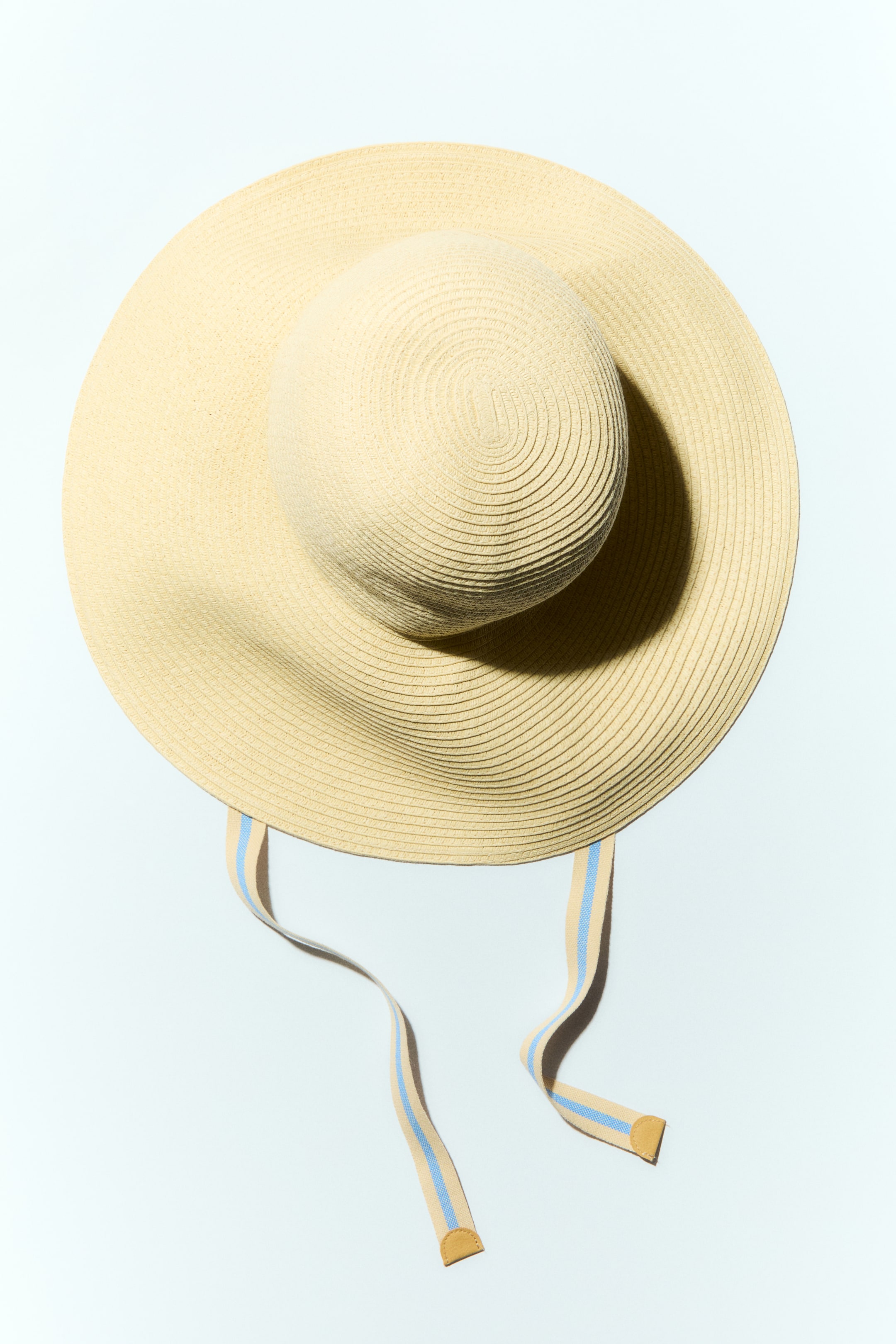 View larger image: A hat crafted from braided paper straw is showcased with a wide brim from a top-down view, featuring cotton-weave chin ties adorned with beige and light blue stripes, extending from the brim and finishing with small brown tabs.