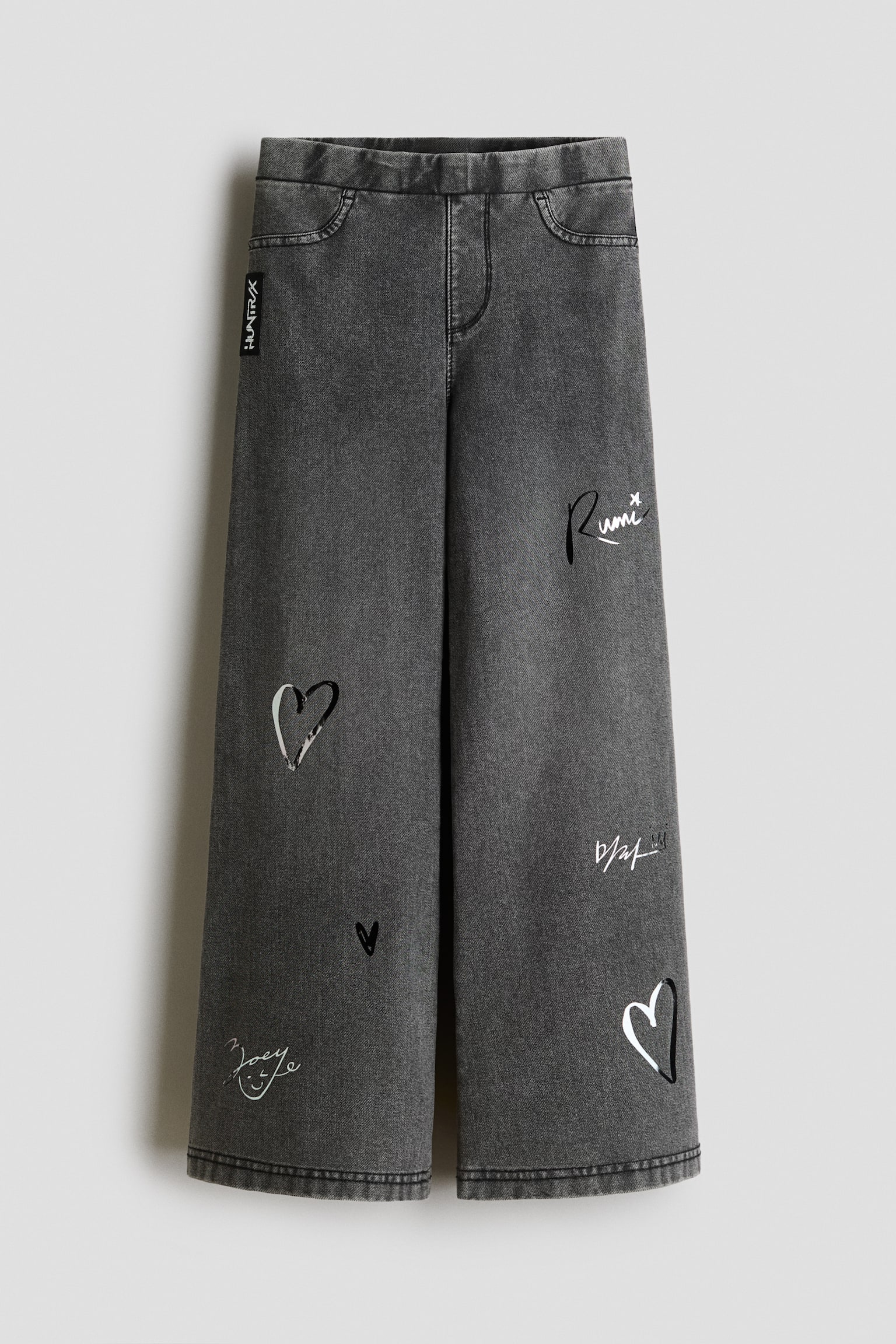These dark grey, denim-look jersey jeggings feature a wide-leg silhouette with an elasticated waistband, mock front pockets and a mock fly. The front of the trousers features printed black-and-white graphics, including outline hearts and handwritten-style script down the legs.