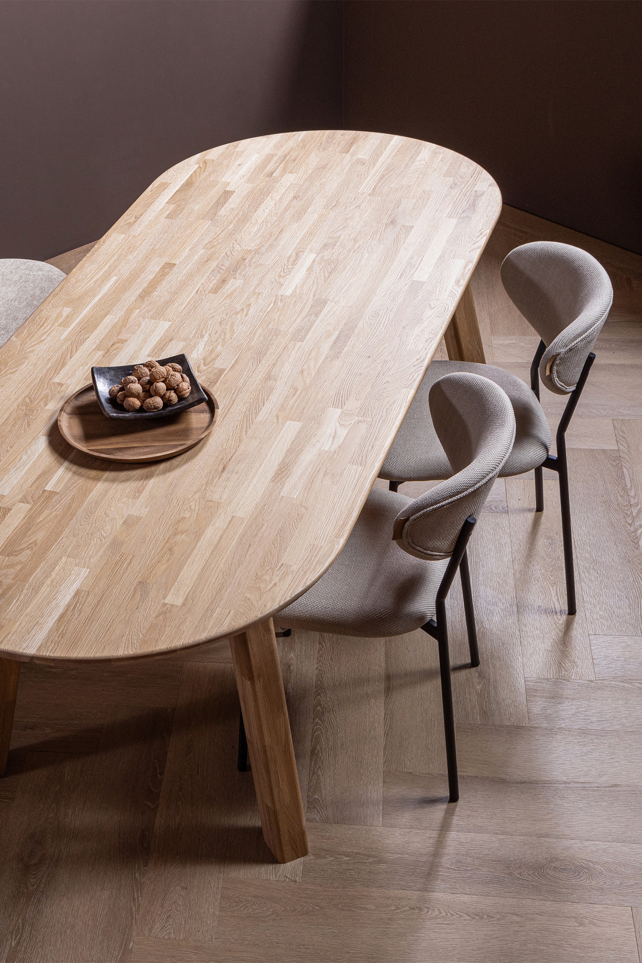 Agrandir l'image: Une table à manger en bois ovale met en valeur sa surface en bois clair à lames, avec un petit bol noir de noix reposant sur un plateau en bois. Trois chaises de salle à manger avec un revêtement en tissu gris clair et des dossiers incurvés sont disposées autour de la table, dotées de cadres en métal foncé.