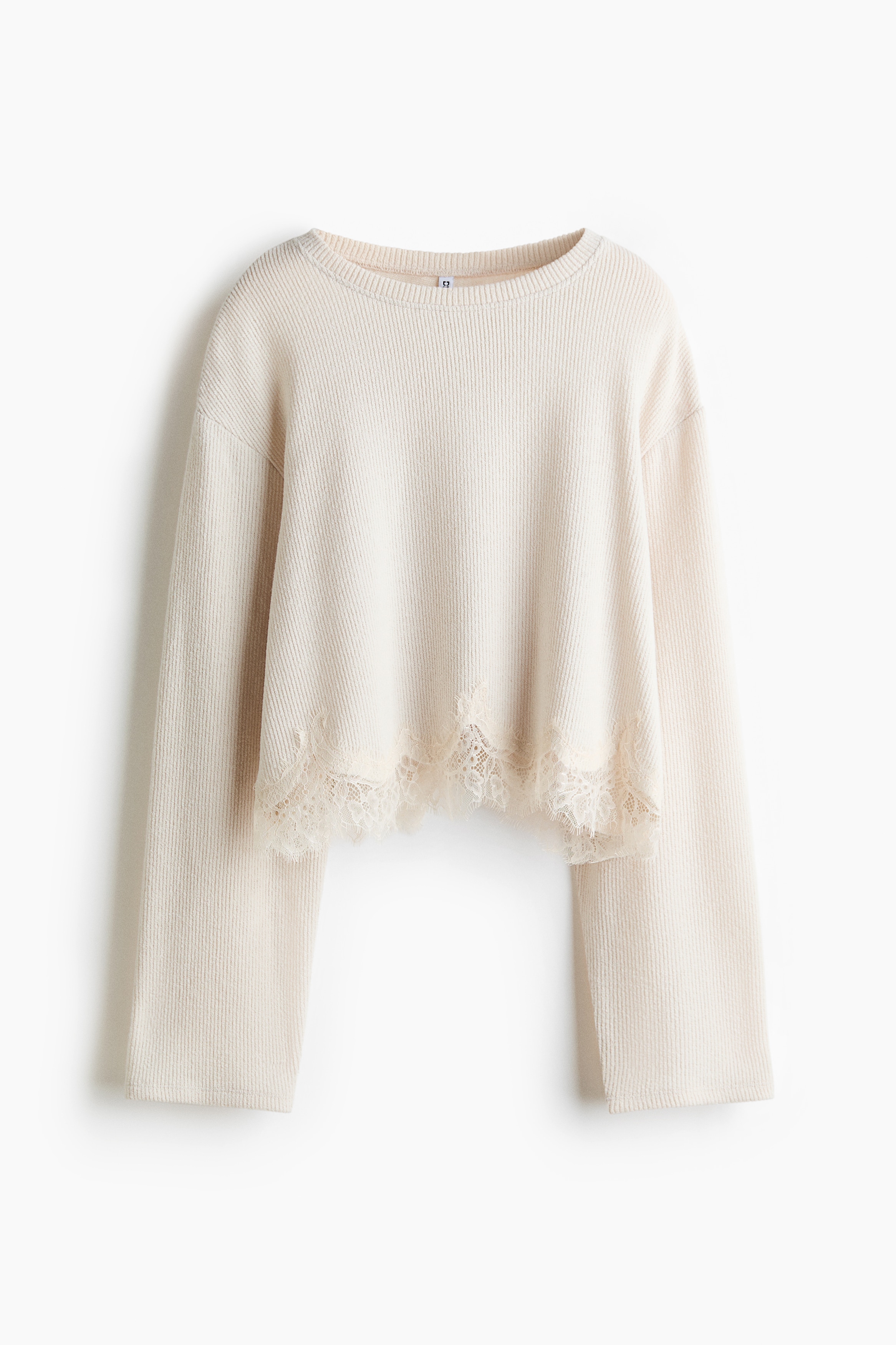 A short, loose-fit knit top in off-white is being worn by the model, featuring a round ribbed neckline, dropped shoulders, long sleeves, and a scalloped, lace-trimmed hem. The top is styled with blue jeans, with the model resting their hands inside the front pockets.