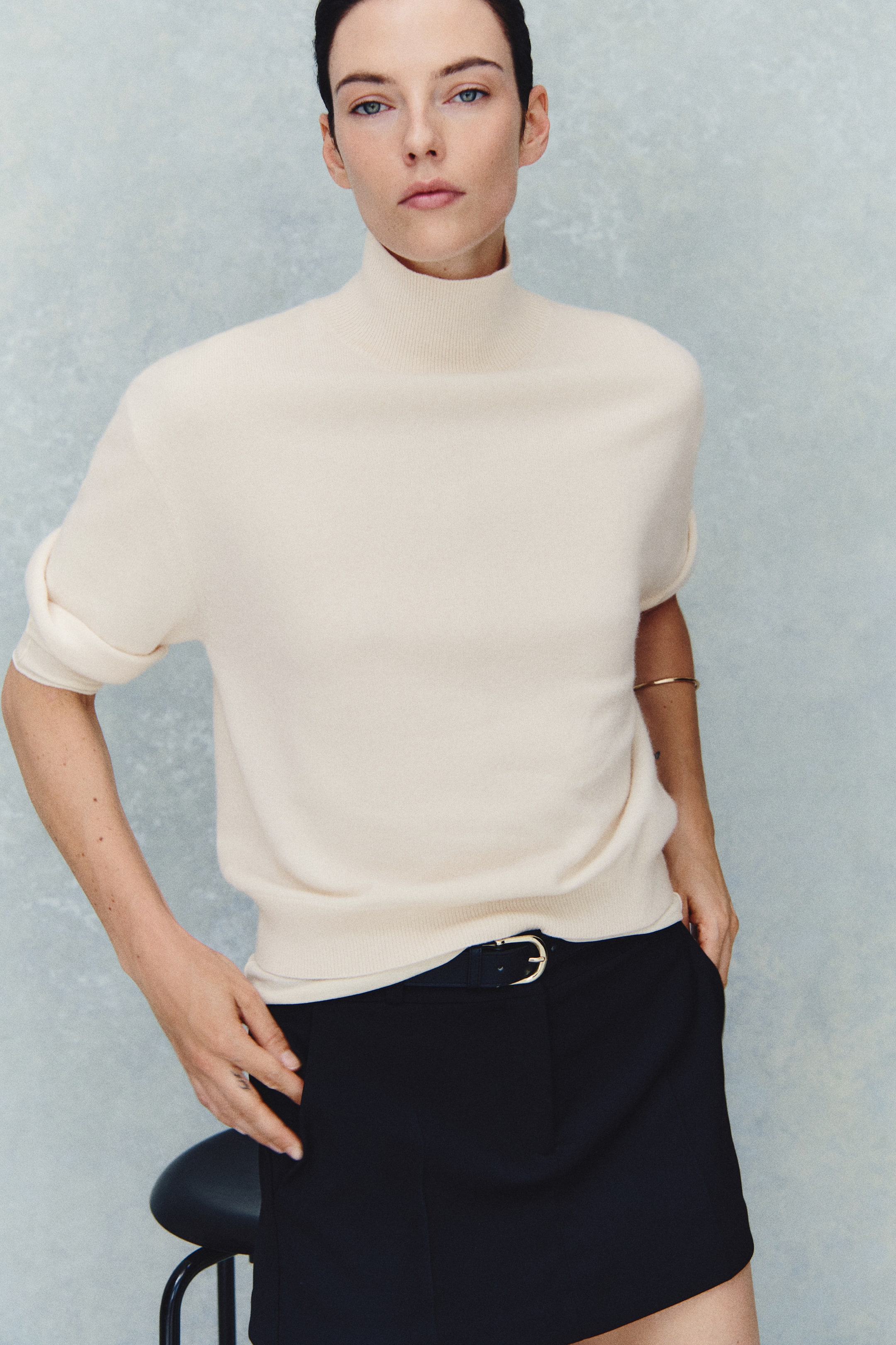 Größeres Bild anzeigen: Ein cremefarbener Feinstrick-Pullover aus Kaschmir mit hohem Rippkragen, überschnittenen Schultern und kurzen Ärmeln wird vom Model getragen. Der Pullover ist in eine schwarze Hose gesteckt, die mit einem schwarzen Gürtel fixiert ist.