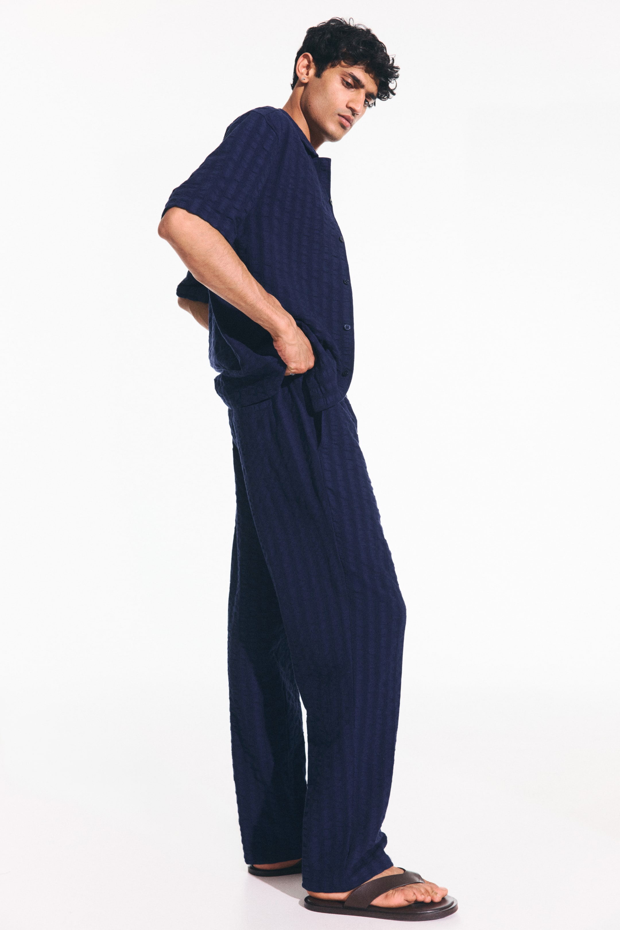 View larger image: The navy blue two-piece set is showcased from a side angle, featuring a short-sleeved top with an open neckline and button-front closure, paired with matching wide-leg pants with side pockets. Both pieces exhibit a subtle textured weave and a relaxed fit.