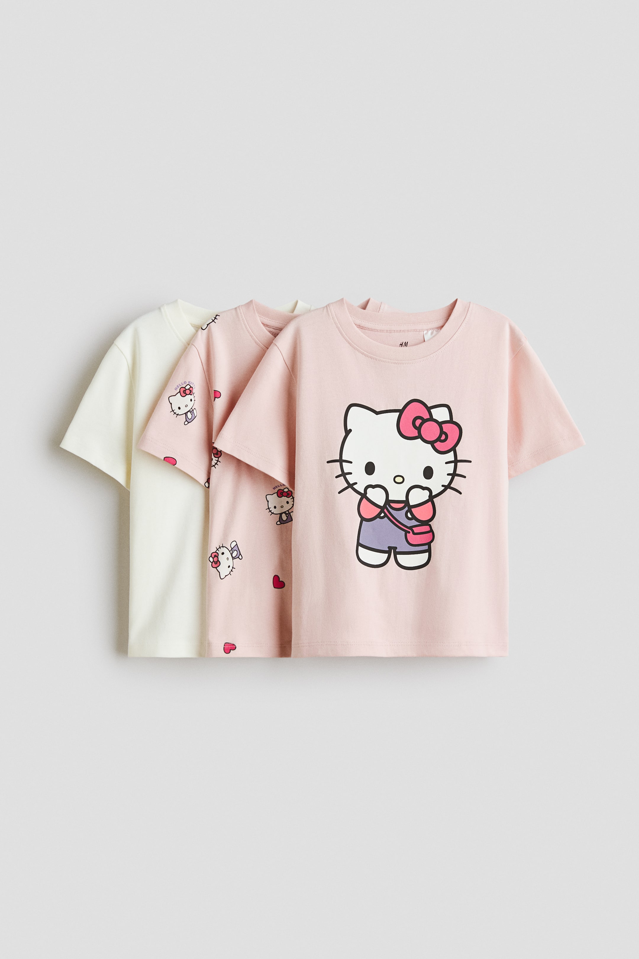 View larger image: Three short-sleeved, crew-neck T-shirts are being presented, layered from left to right, all facing forward. The leftmost T-shirt is cream, the middle T-shirt is light pink with small Hello Kitty patterns and hearts, and the rightmost T-shirt is light pink featuring a large Hello Kitty graphic.