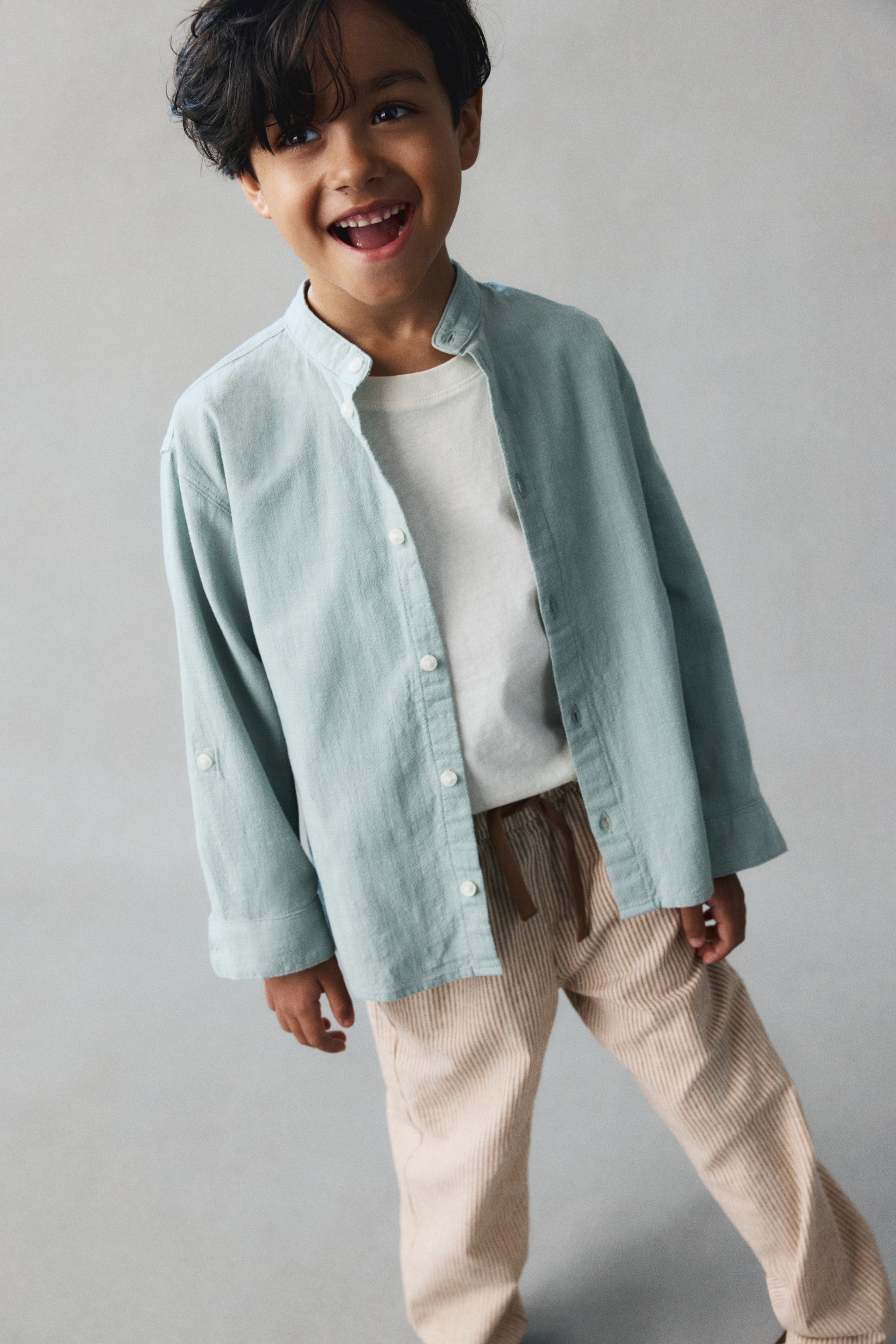 View larger image: Pants in a beige and white striped cotton-linen blend are shown from the front, featuring a relaxed fit, an elasticated waistband, and a contrasting brown drawstring. The pants are styled with a tucked-in white T-shirt and an open light green shirt that frames the waistband and drawstring detail.