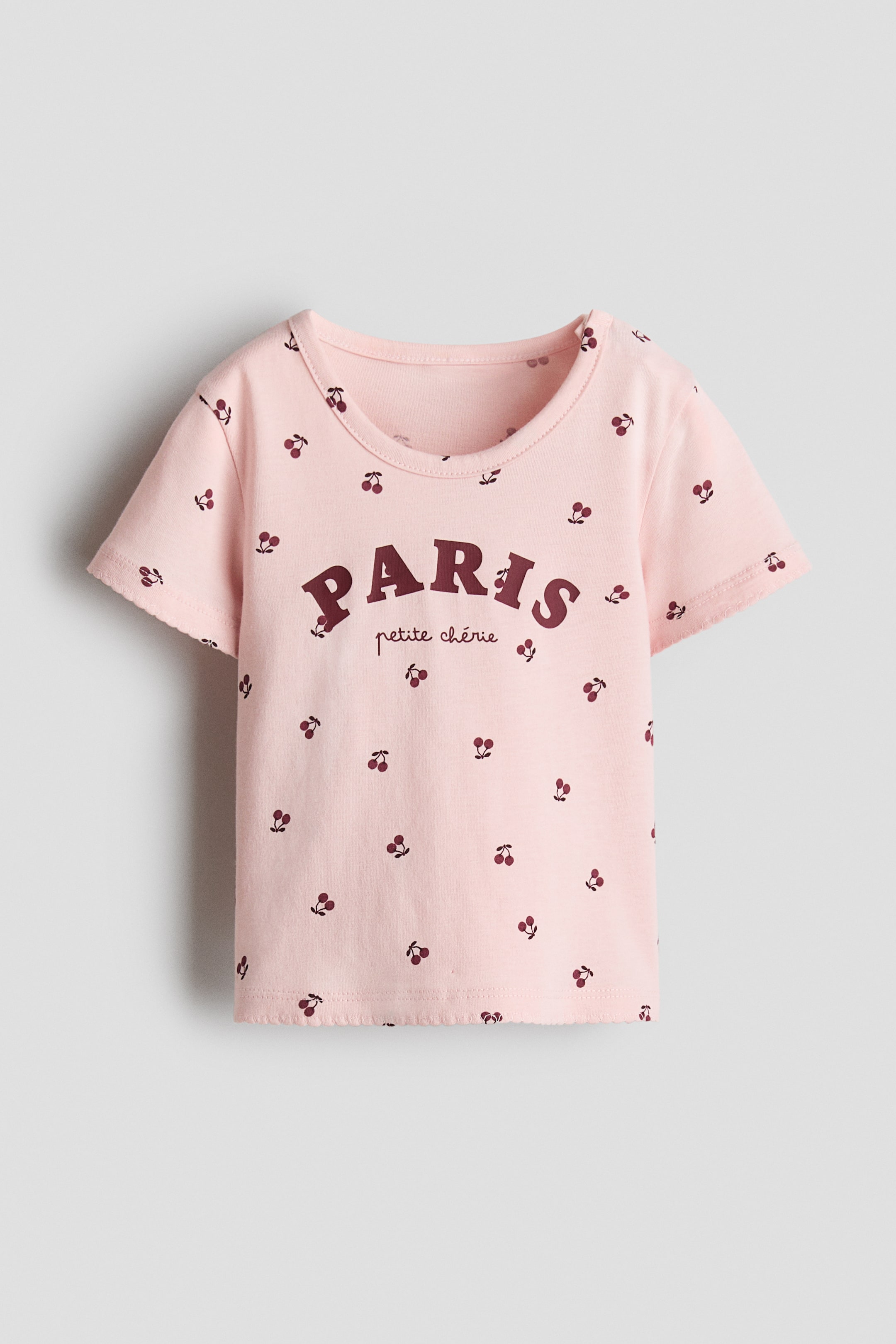 View larger image: The top is shown from the front, made from soft pink cotton jersey and featuring a scattered dark red cherry print. It is designed with a round trimmed neckline and short sleeves, with "PARIS petite ch�rie" printed in dark red across the chest.