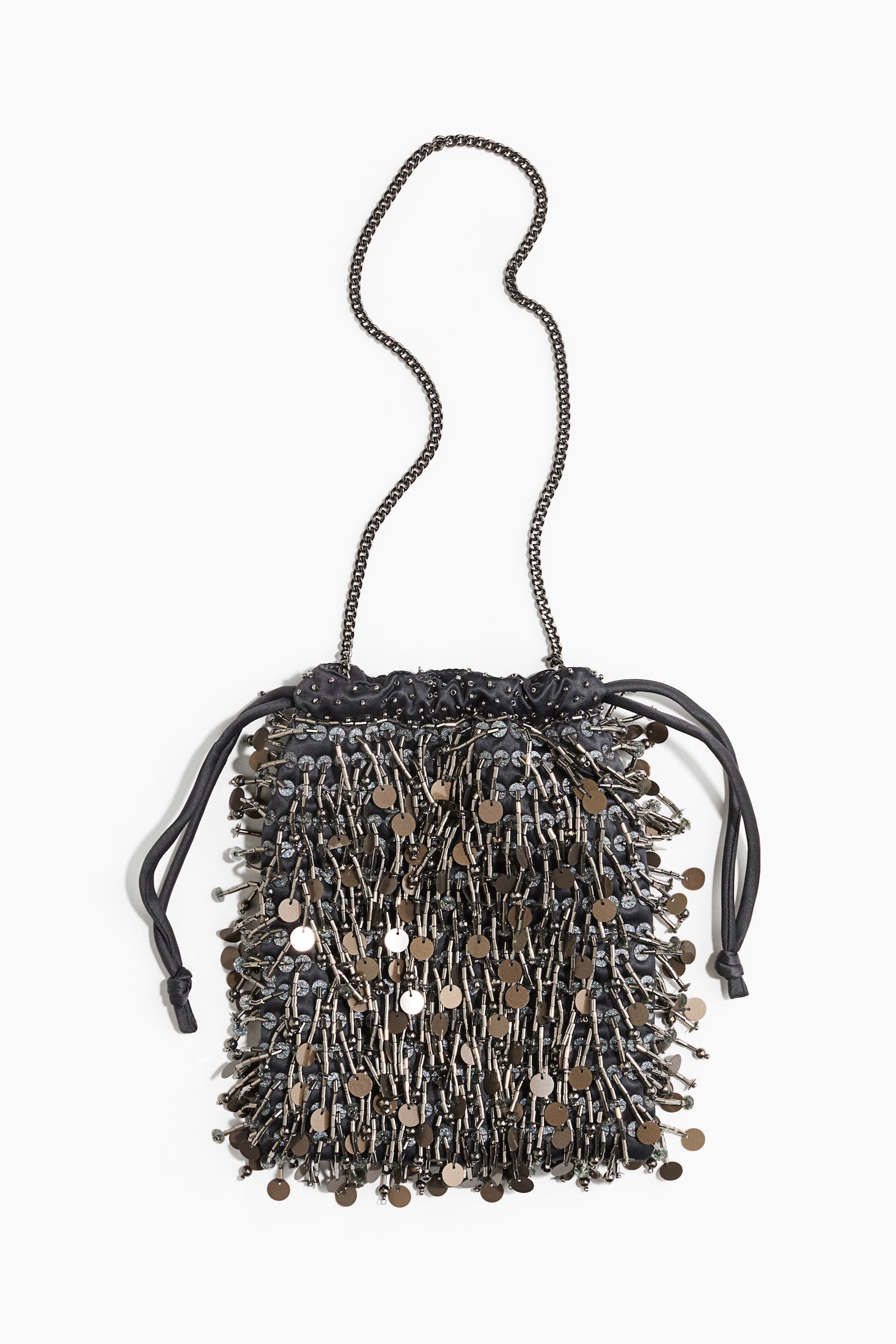 A black drawstring pouch bag is facing front, displaying a dense layer of dangling gunmetal-toned bugle beads and flat, round metallic sequins. The bag is featuring a thin metal chain shoulder strap and black fabric drawstring cords knotted at the sides.