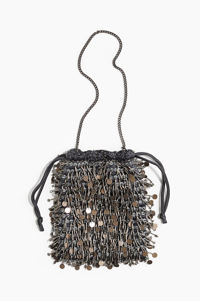 Una bolsa de cordón negra aparece de frente, mostrando una densa capa de cuentas tubulares colgantes en tono gunmetal y lentejuelas metálicas planas y redondas. La bolsa presenta una fina correa de cadena metálica para el hombro y cordones de tela negros con nudo en los laterales. - 1