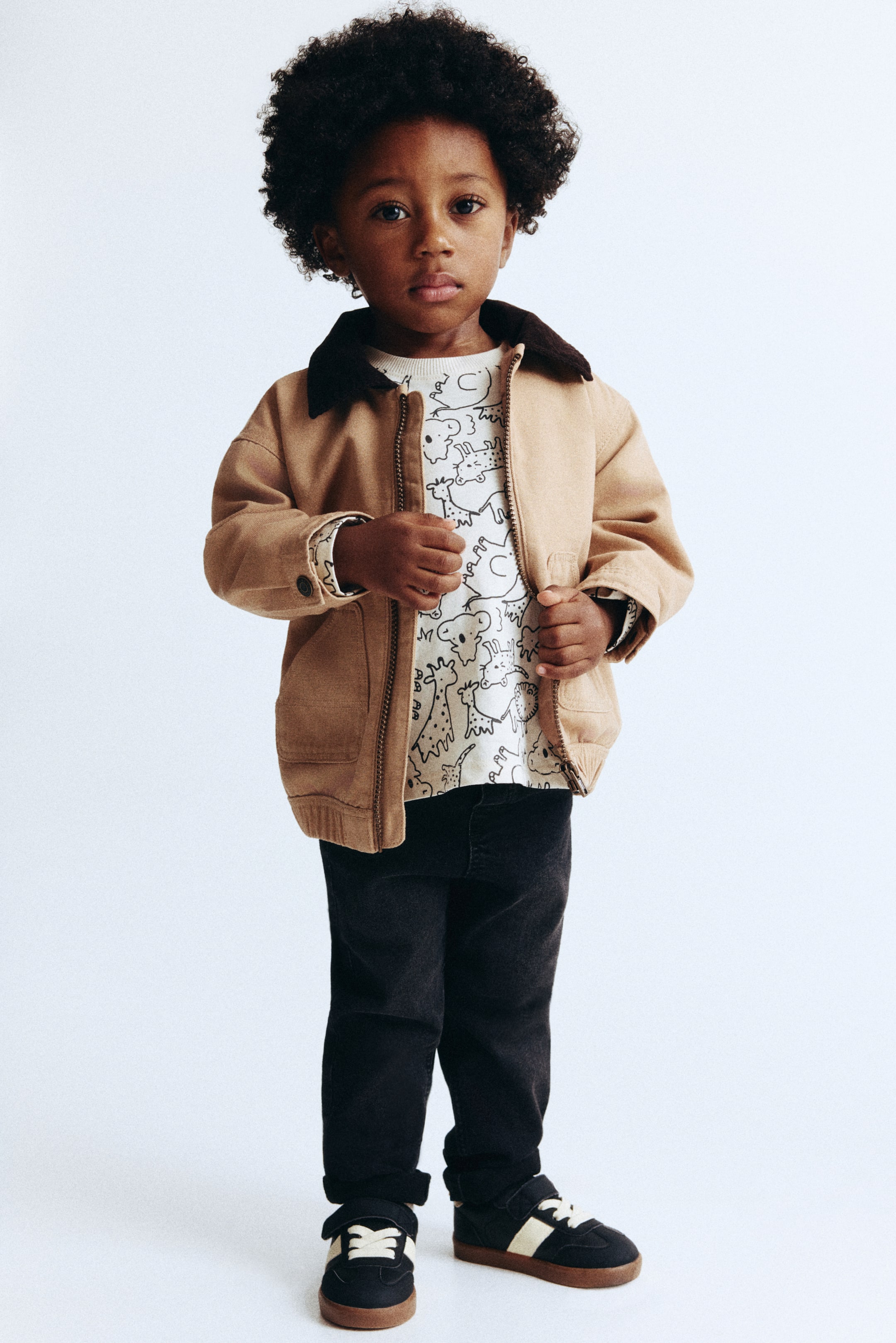 View larger image: This kids’ outfit features a tan utility jacket with a dark brown corduroy collar, full front zip, and ribbed cuffs and hem, shown facing forward. A cream long-sleeved top with black line-art animal print is layered underneath, while dark grey corduroy trousers and black sneakers with beige hook-and-loop straps and gum soles complete the look.