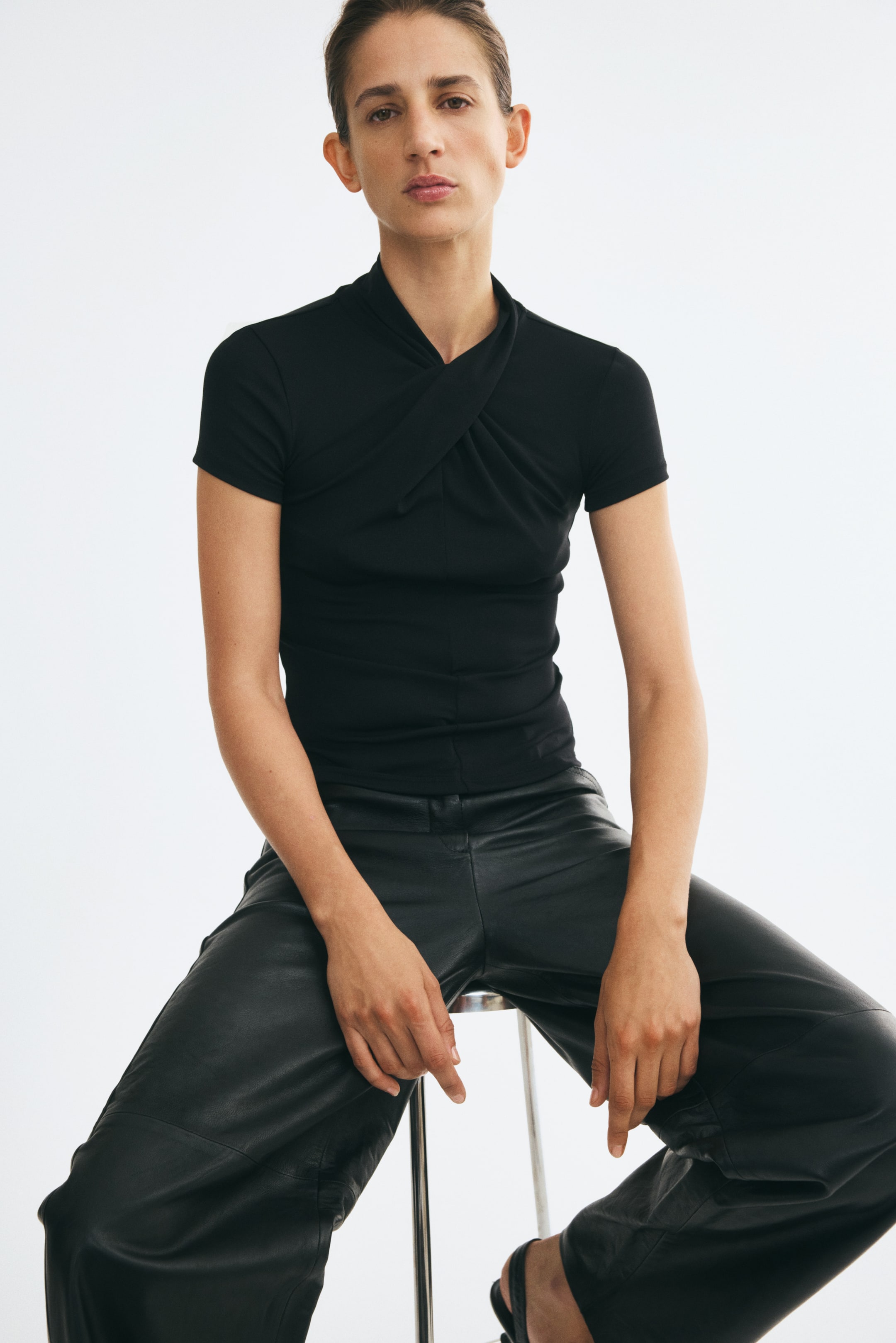 View larger image: The model is wearing a black, short-sleeved fitted top featuring a stand collar with a twisted, draped detail at the front. The top is styled with wide-leg black leather trousers.