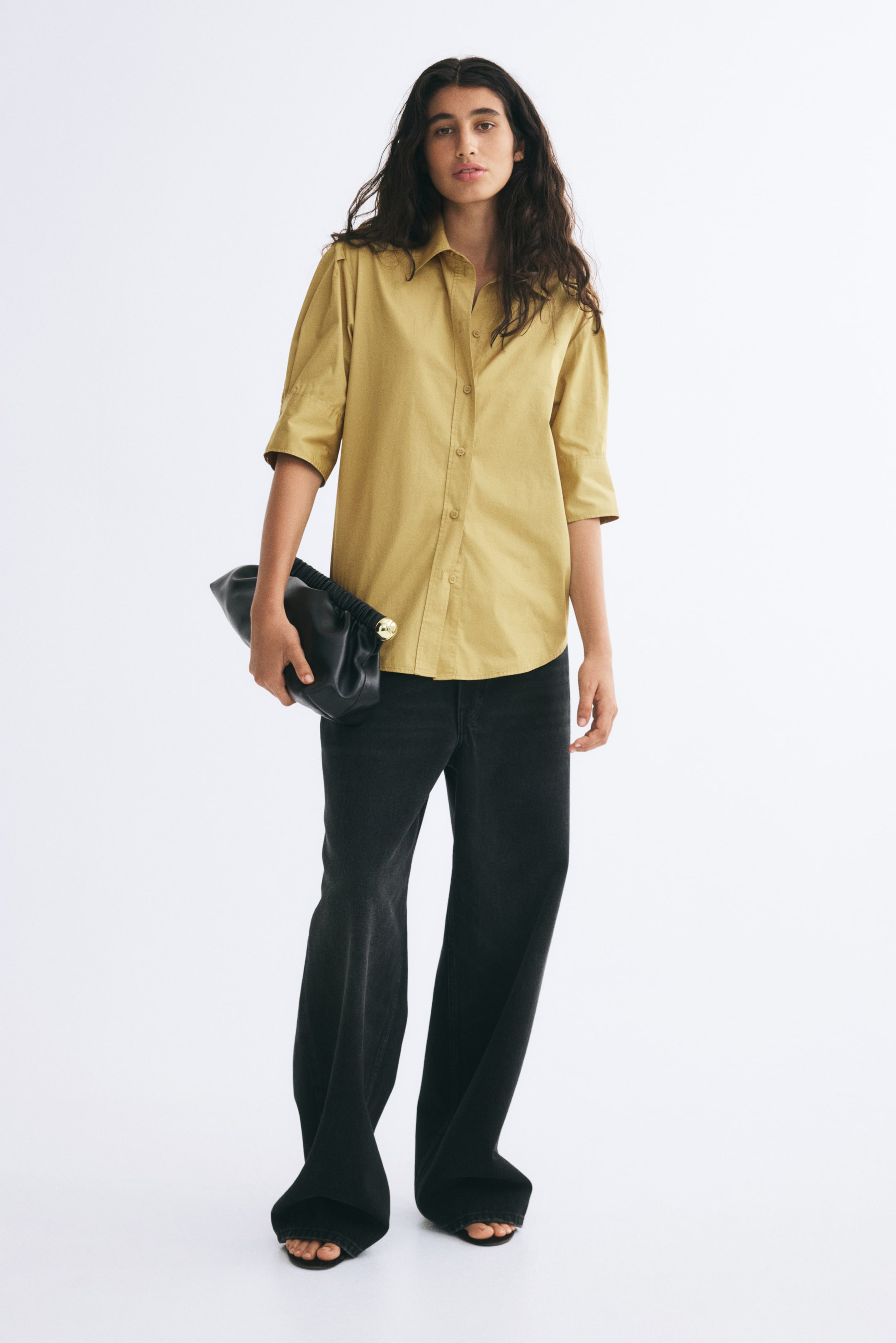 View larger image: A mustard-yellow cotton poplin shirt featuring a collar, a button-down front, and elbow-length pleated sleeves is being worn by the model. The model is styling the untucked shirt with black wide-leg trousers while holding a black clutch bag.