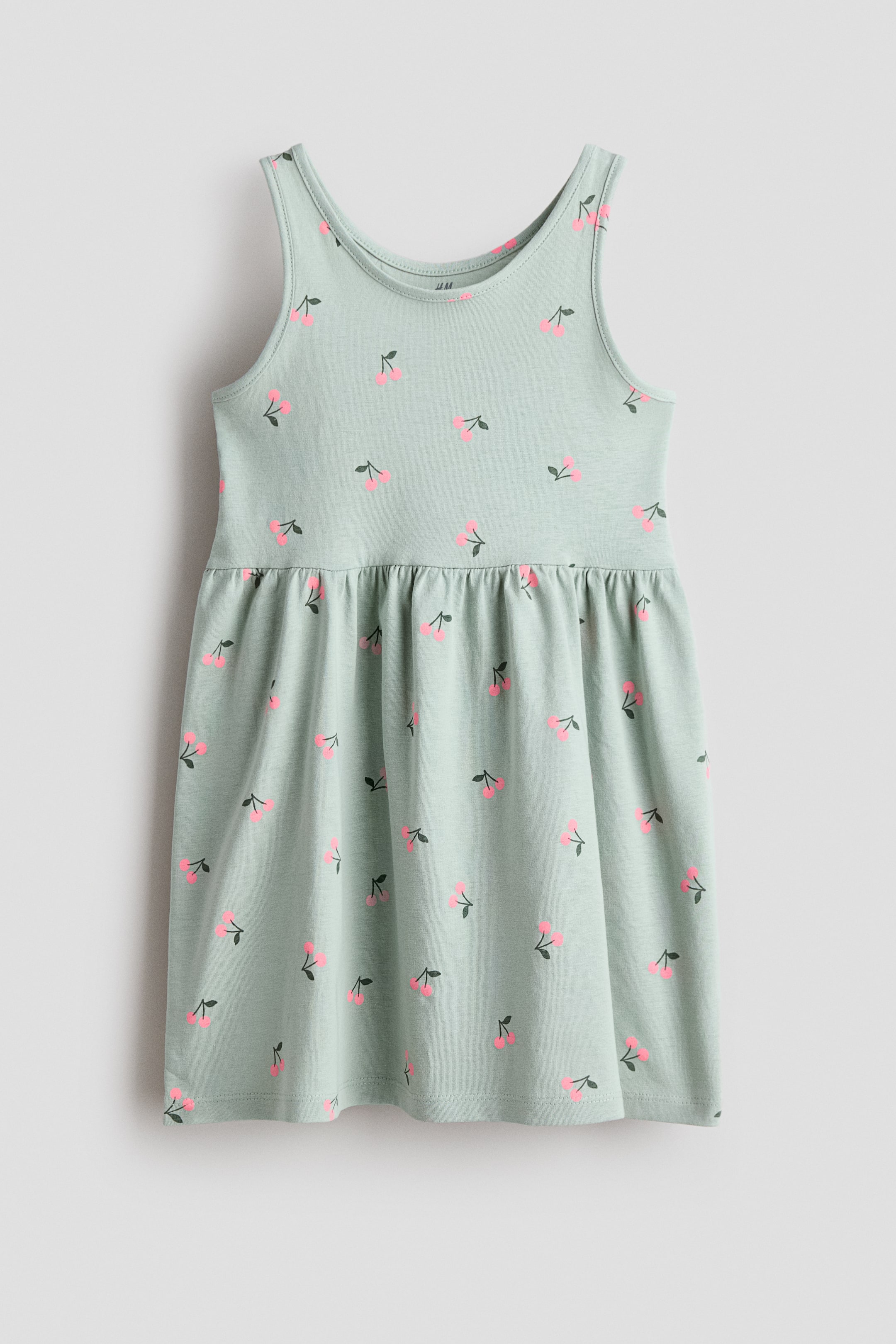 Größeres Bild ansehen: Ein ärmelloses Kleid zeigt seine FRONT, gefertigt aus weichem, hellgrünem Baumwolljersey und mit einem rosa Kirschenmuster. Es ist mit einer in der Taille gerafften Naht und einem sanft ausgestellten Rock gestaltet.
