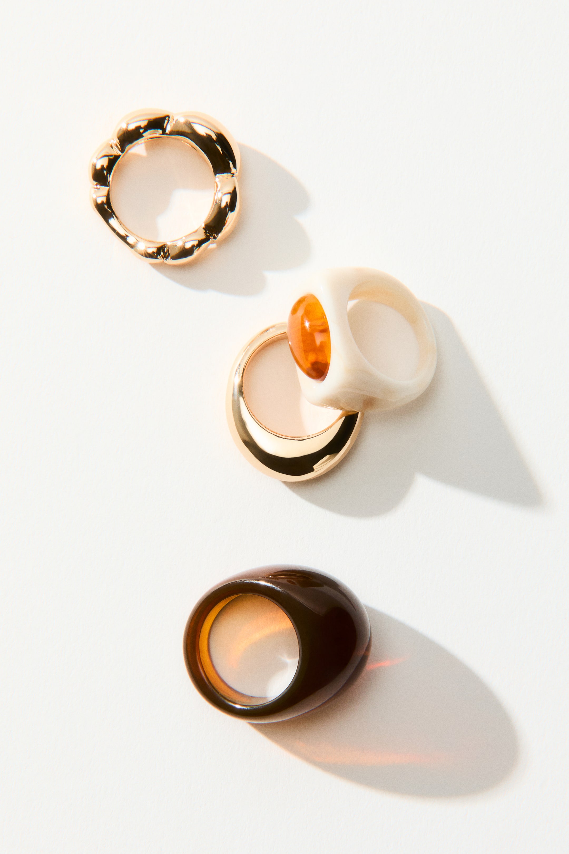 View larger image: Rings are showcasing a variety of designs in metal and resin, all laid flat. At the top, there's a chunky, wavy gold-toned metal ring. In the center, a smooth gold-toned metal band is layered with a cream-colored resin ring, featuring an amber resin gem. At the bottom, a dark brown, domed resin ring is also displayed.
