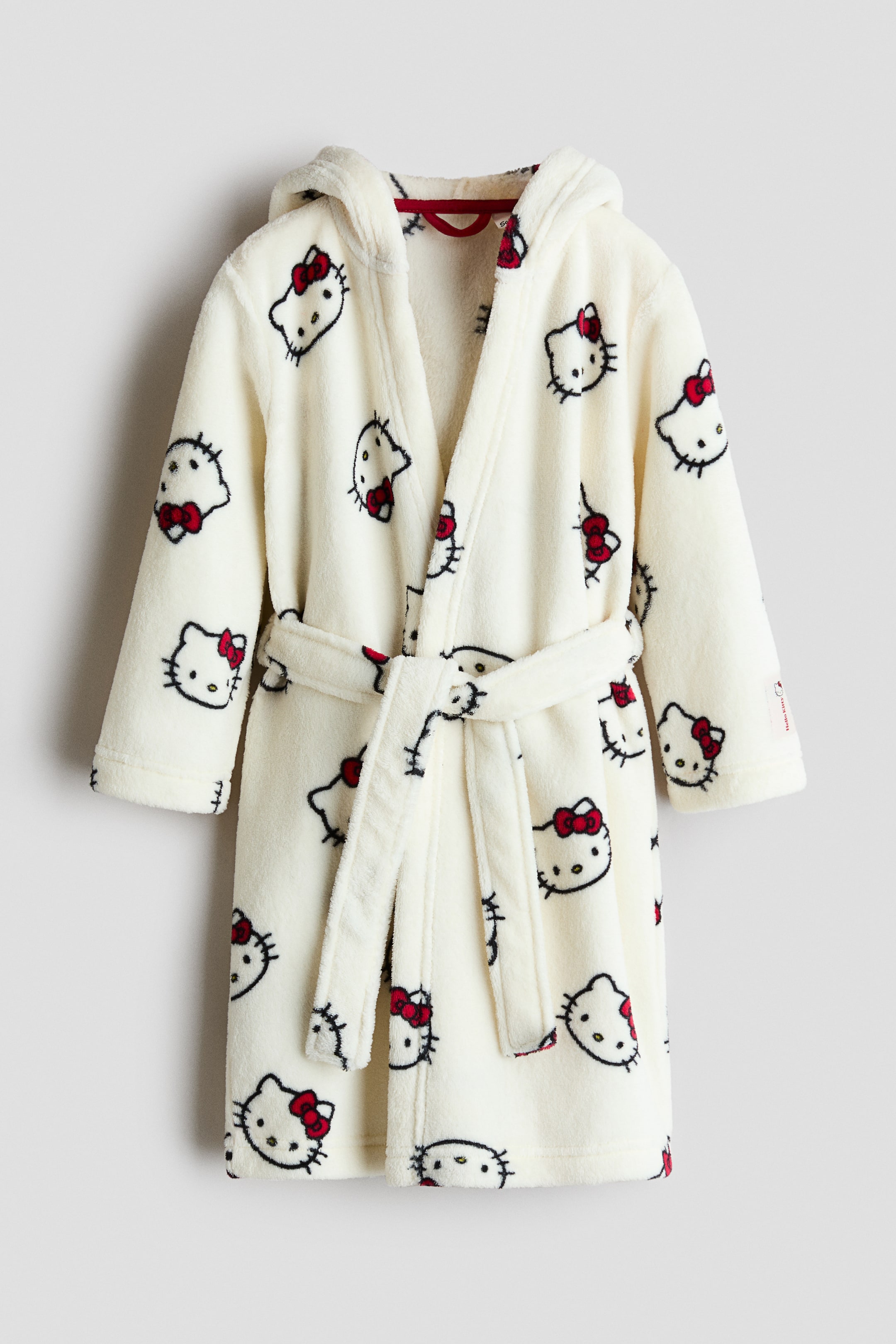 View larger image: A cream fleece dressing gown is shown from the front, featuring an attached hood and an all-over pattern of Hello Kitty faces with red bows. A matching detachable tie belt cinches the waist.