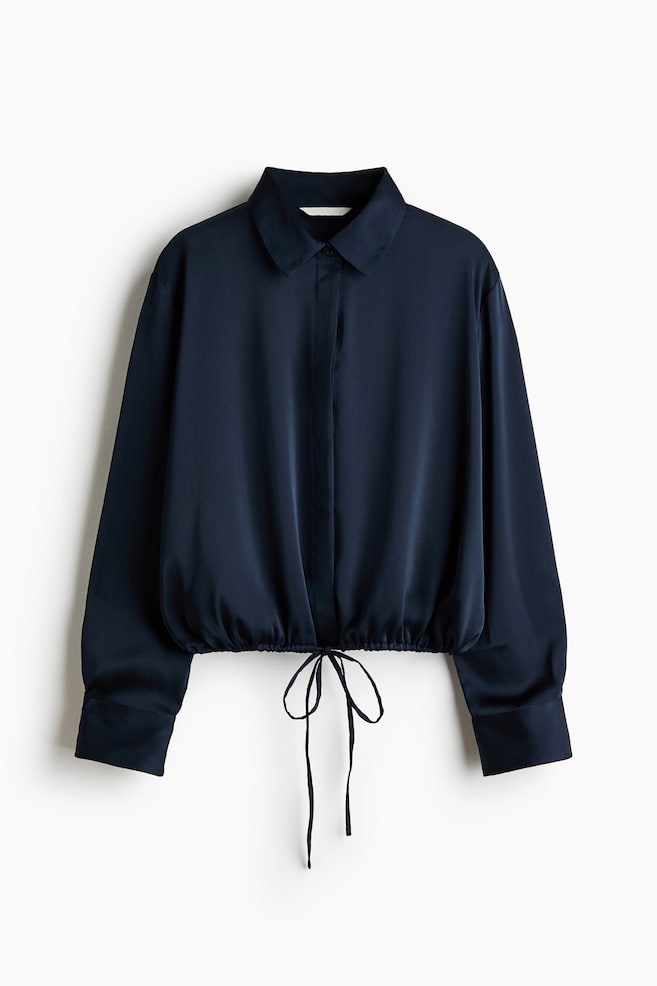 Uma camisa de cetim azul-marinho escuro está pendurada, voltada para a frente, apresentando uma gola clássica, carcela de botões oculta e mangas compridas com punhos largos com botões. A bainha inferior está franzida com um cordão ajustável, atado em laço ao centro. - 1