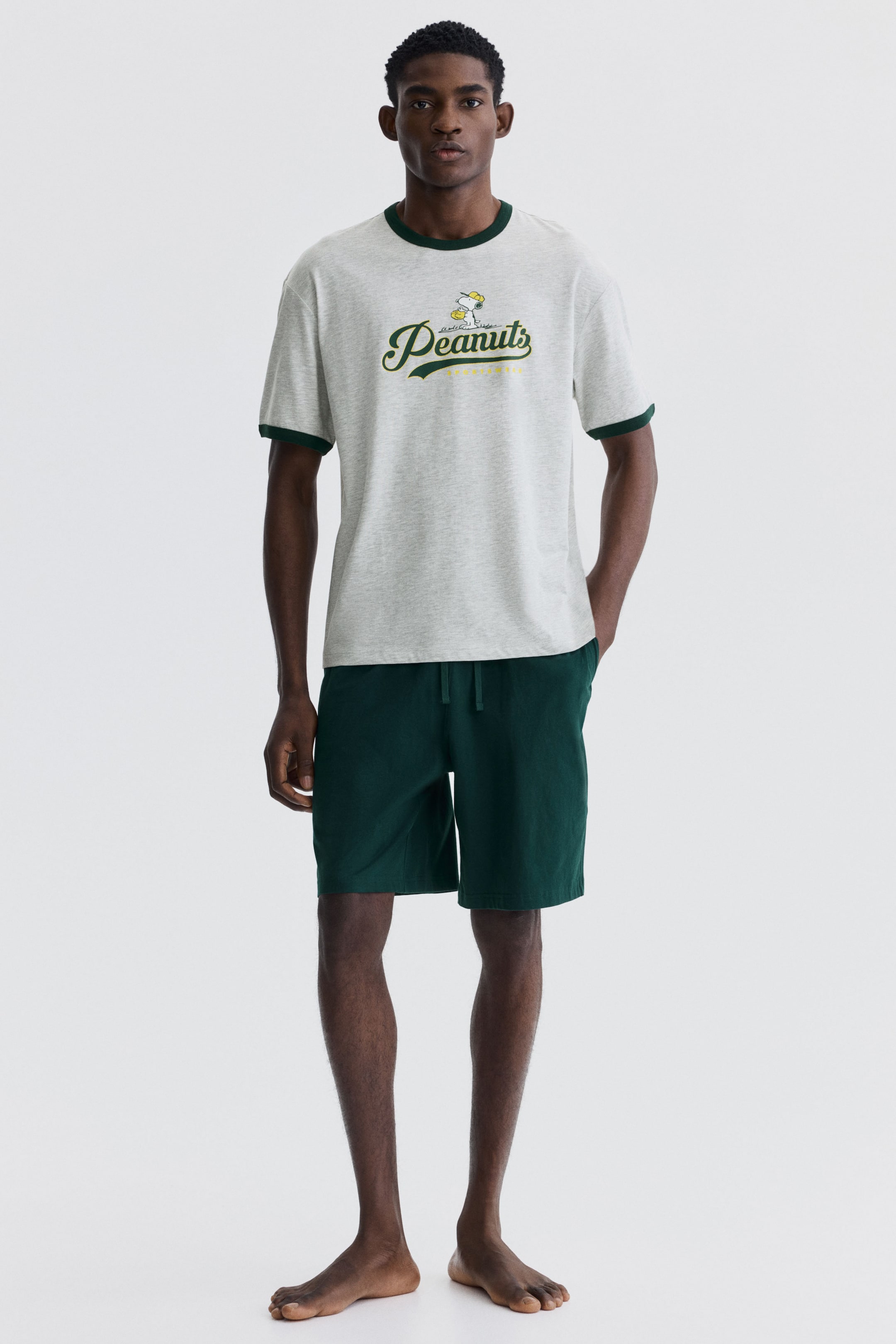 Grotere afbeelding bekijken: Een tweedelige loungewear set wordt gepresenteerd vanuit een frontale hoek. Het lichtgrijze marl t-shirt heeft een donkergroene geribbelde ronde hals en bijpassende korte mouwboorden, met "Peanuts" tekst en een Snoopy karakterontwerp op de borst. De donkergroene shorts hebben een elastische taille met trekkoord en reiken tot halverwege de dij.