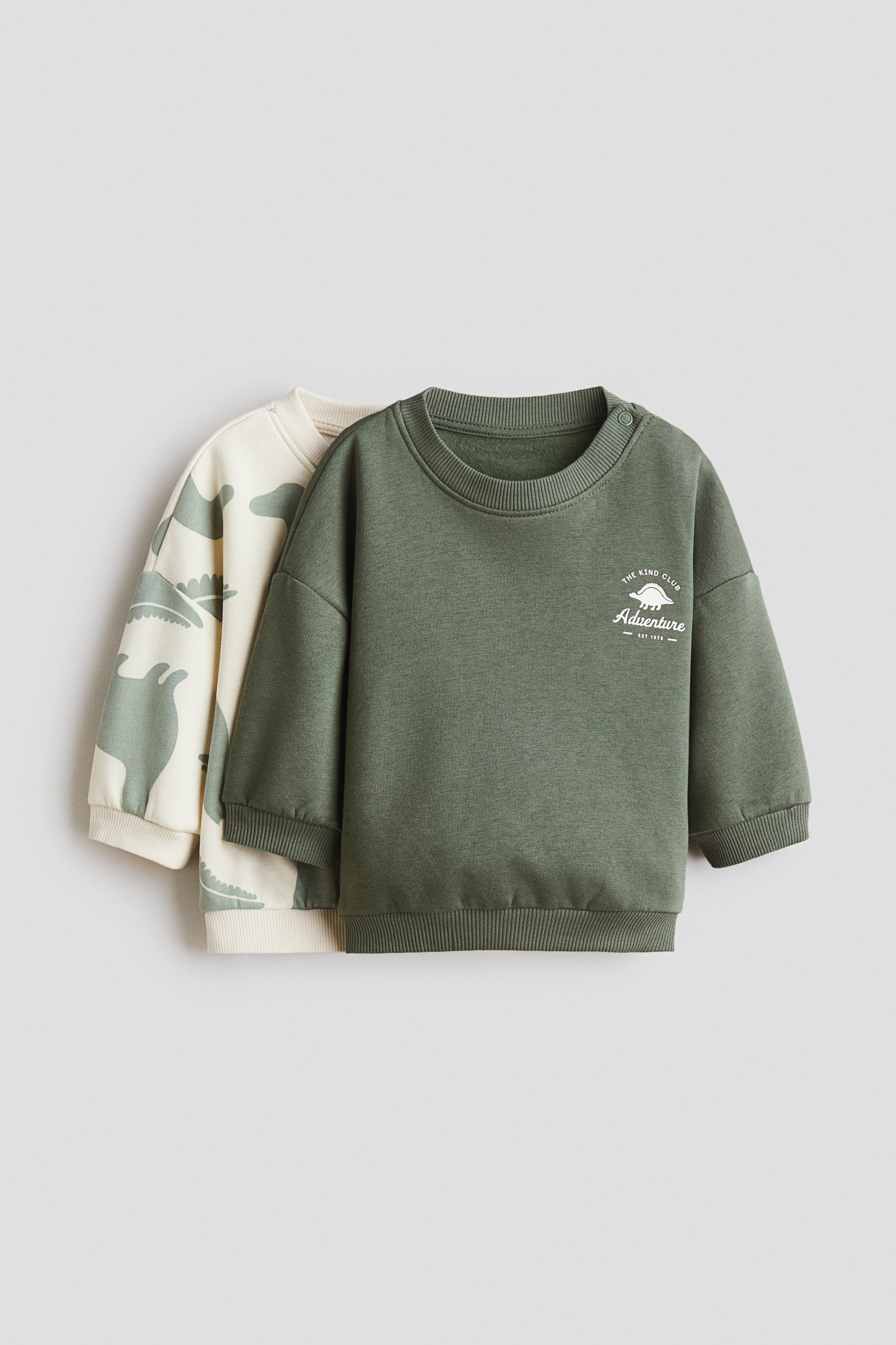 View larger image: Two baby sweatshirts are laid flat, facing forward. The olive green sweatshirt at the front features a white “Adventure” graphic with a small dinosaur icon on the chest and two snap buttons on the left shoulder. The off-white sweatshirt, layered behind, is patterned with olive green dinosaurs. Both sweatshirts are finished with ribbed necklines, cuffs, and hems.