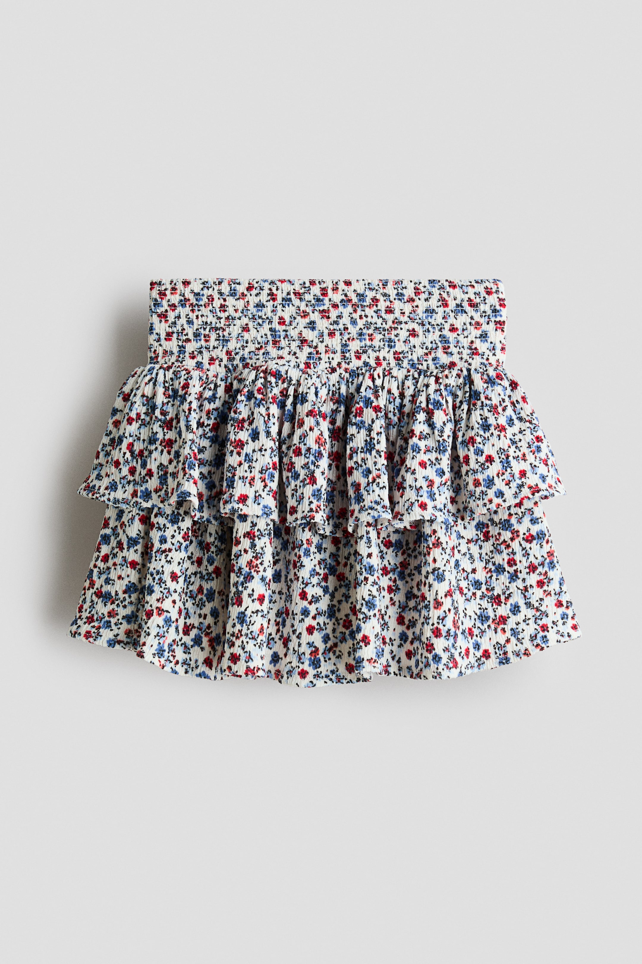 View larger image: A skort in crinkled jersey is displaying its front, featuring an all-over floral print of small red and blue flowers on a white background. It has a wide, smocked waist panel and two ruffled tiers forming the skirt.