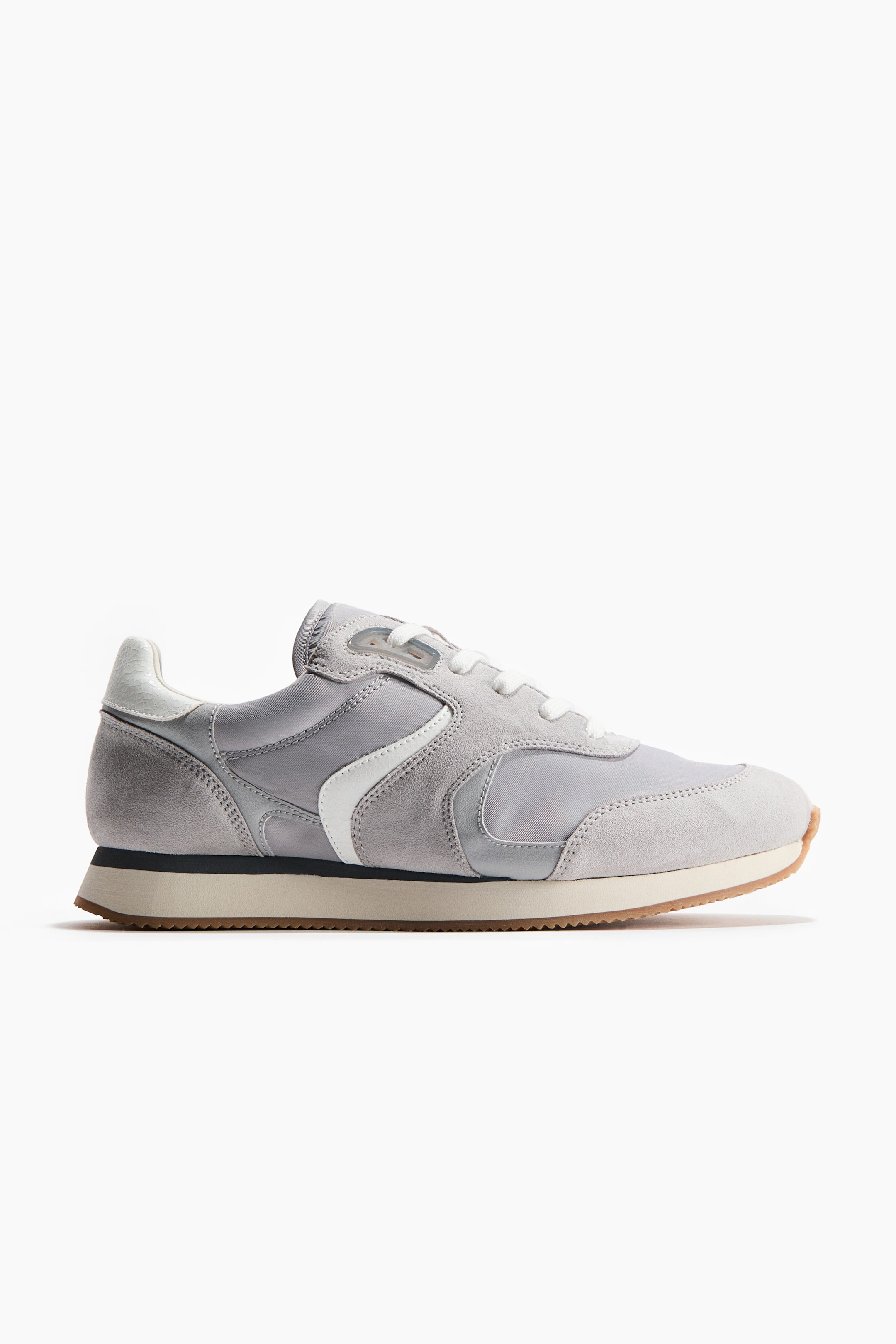 View larger image: A light gray sneaker is showcasing its side profile, featuring a paneled upper of light gray suede and fabric with white laces. A white curved stripe adorns the side, and the sole consists of an off-white midsole with a brown rubber outsole.