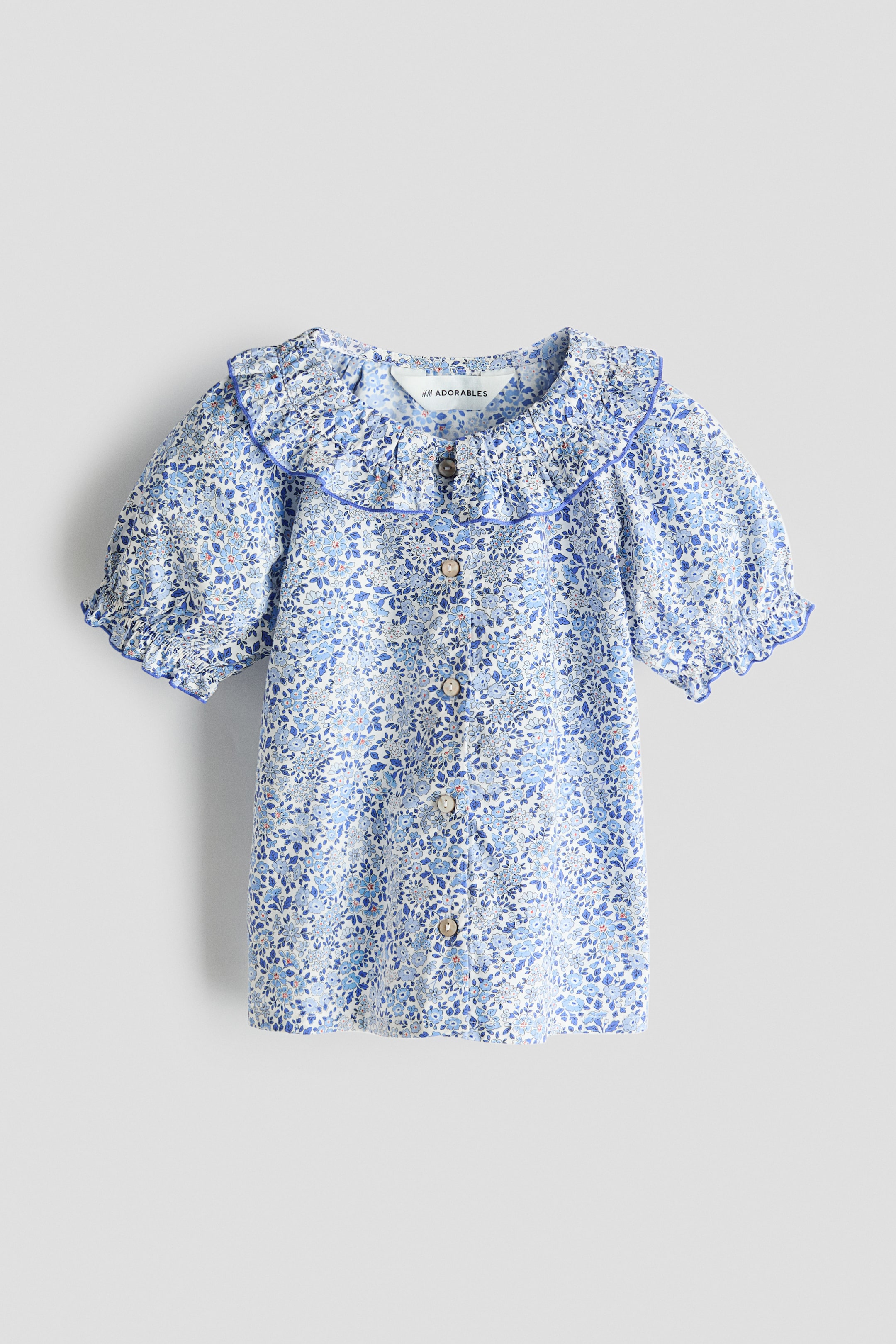 Ampliar la imagen: Una blusa de manga corta para niña aparece de frente, mostrando un tejido blanco con un patrón floral azul en toda la superficie. Presenta un cuello con volantes y ribetes azules, mangas cortas abullonadas con puños con volantes y ribetes, y un cierre de botones completo al frente con botones redondos de color marrón claro.