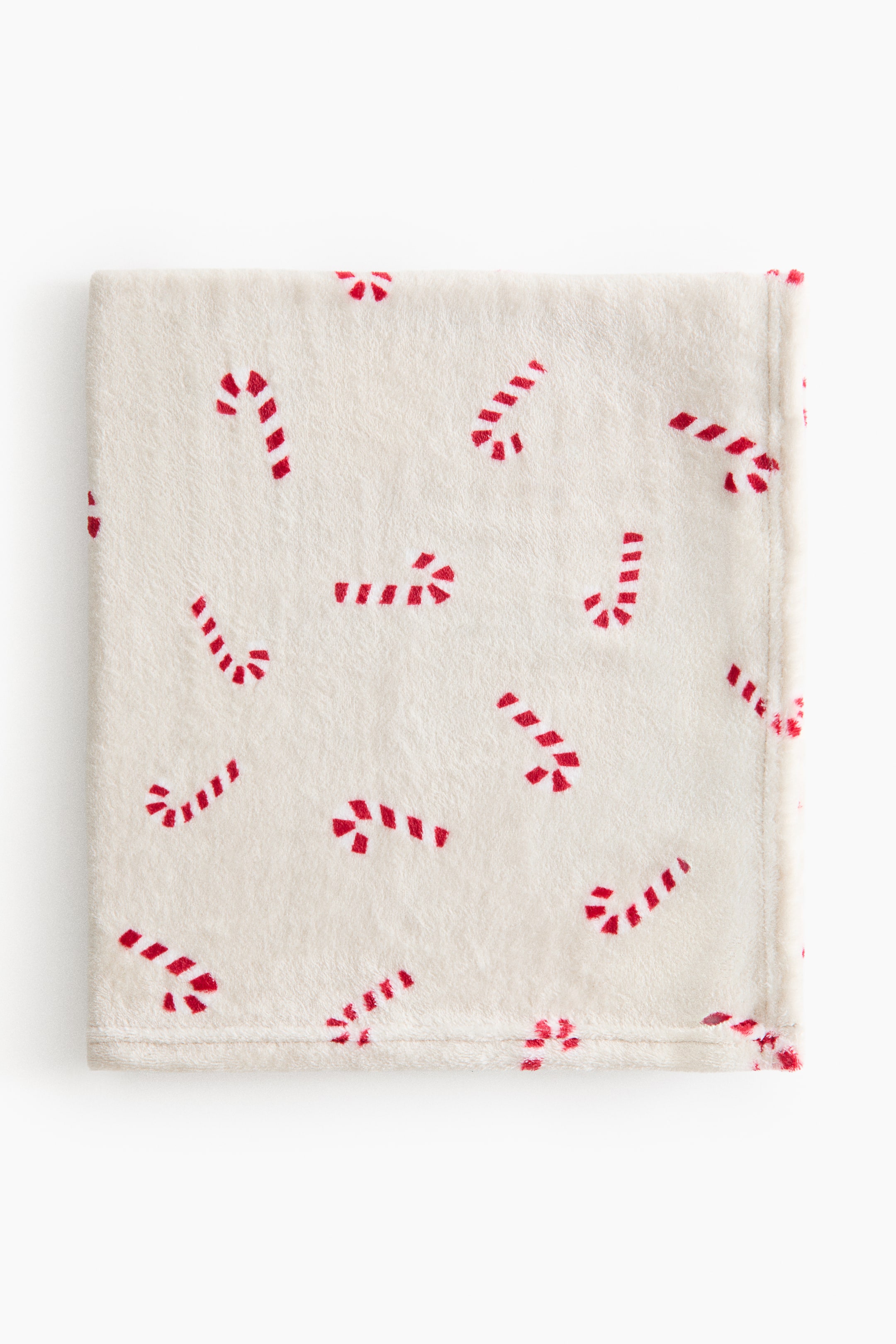 Ampliar la imagen: Una manta para bebé está hecha de suave tela polar color crema, con un estampado total de bastones de caramelo a rayas rojas y blancas dispersos por toda la superficie. Está doblada en un rectángulo ordenado y se presenta en una vista desde arriba.