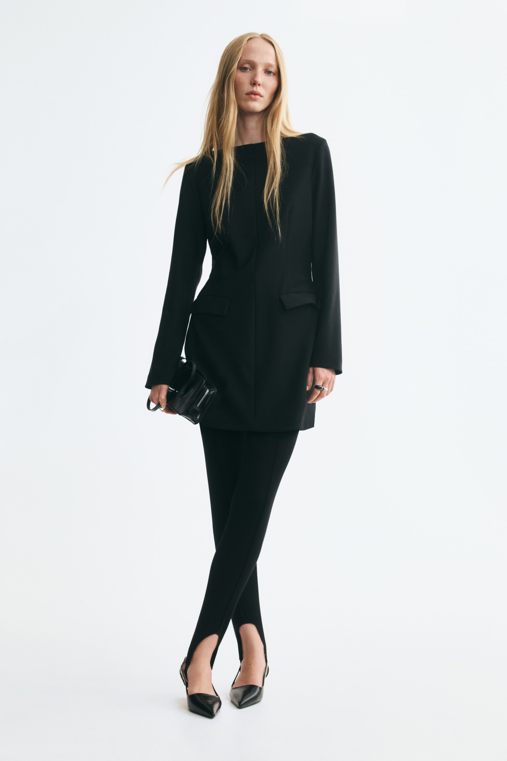 View larger image: A short black stretch-jersey dress with a boat neckline, long sleeves, waist darts and front flap pockets is worn by the model. The outfit is styled with black stirrup leggings and pointed-toe heels, with a small black clutch held in one hand.