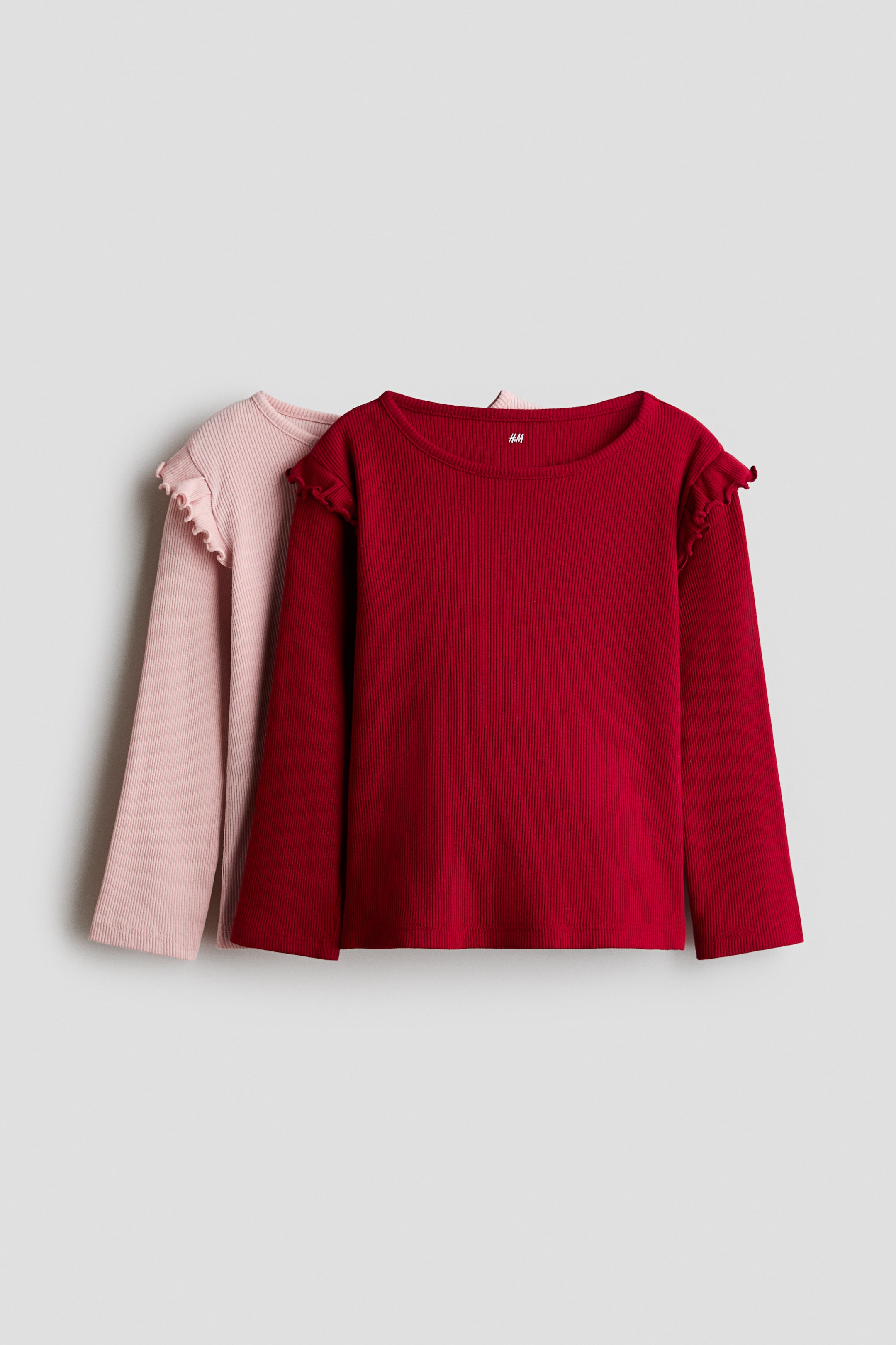 View larger image: Two long-sleeved ribbed jersey tops are shown from the front, with a dark red top overlapping a light pink top. Both tops feature a round neckline with narrow trim and a ruffled flounce over the shoulders.
