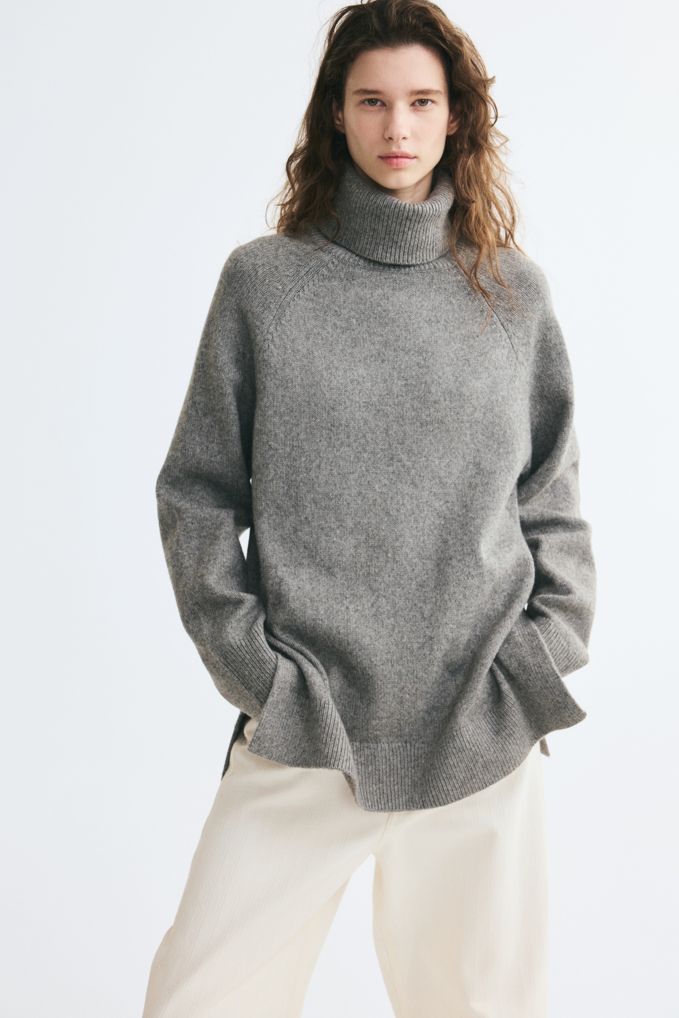 A grey oversized jumper in a soft knit is shown from the front, featuring a ribbed polo neck and long raglan sleeves with ribbed cuffs. The relaxed silhouette falls to a ribbed hem with a small slit at each side.