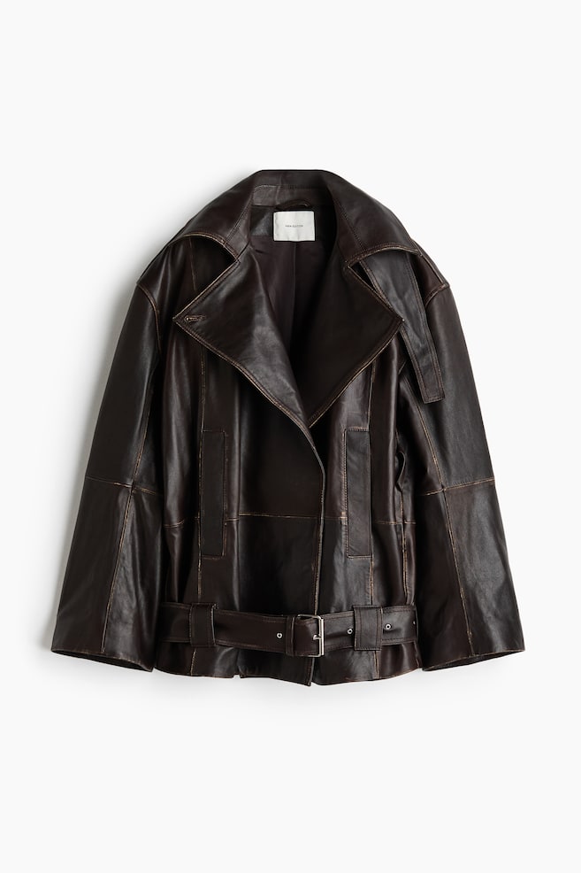 A dark brown leather jacket is hanging facing the front, featuring an oversized silhouette with wide, cropped sleeves and large notched lapels. The jacket displays distressed seam details, vertical welt pockets, and a matching leather belt with a silver-tone buckle at the hem. - 1