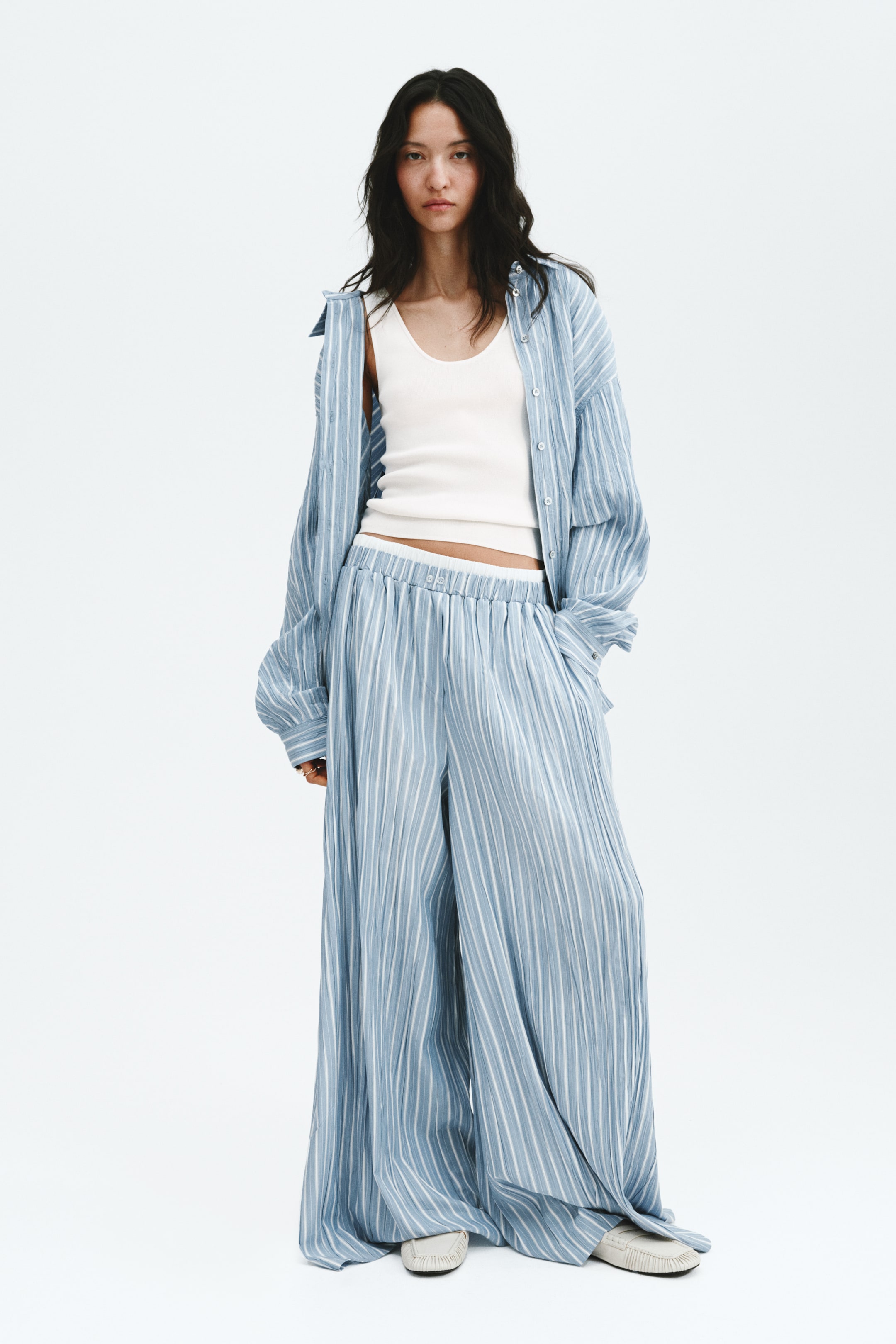 Agrandir l'image: Une tenue coordonnée à rayures verticales bleu clair et blanc est présentée, comprenant une chemise boutonnée oversize portée ouverte sur un débardeur blanc côtelé à col rond. La tenue est complétée par un pantalon large plissé assorti, doté d'une taille élastiquée, le tout présenté de face.