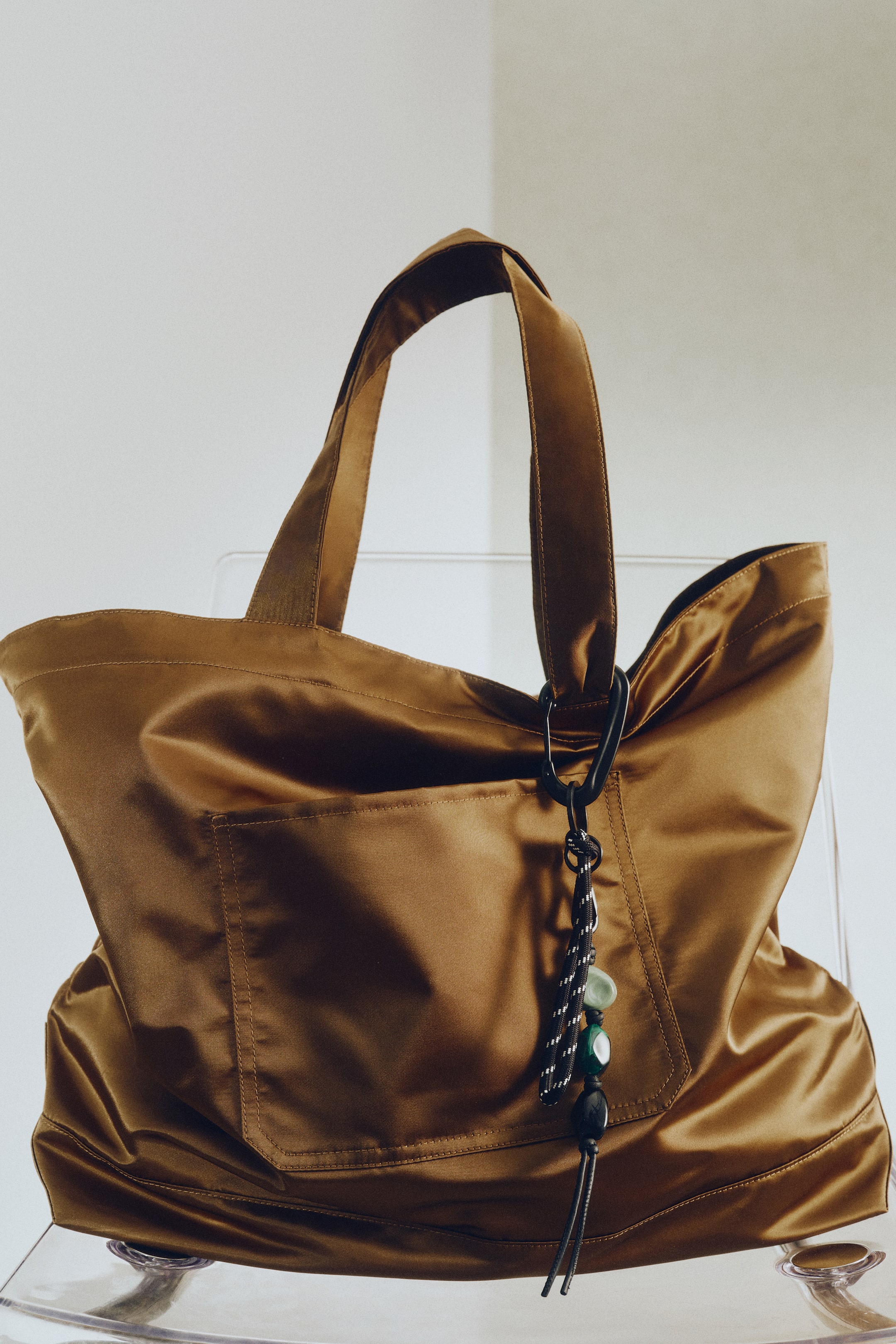 View larger image: A golden brown satin-like tote bag is being presented at a slight angle, showcasing its soft, unstructured form. It features two wide shoulder straps and an exterior patch pocket on its front. A decorative black carabiner clip holds a braided cord charm with green and black beads from one of its straps.