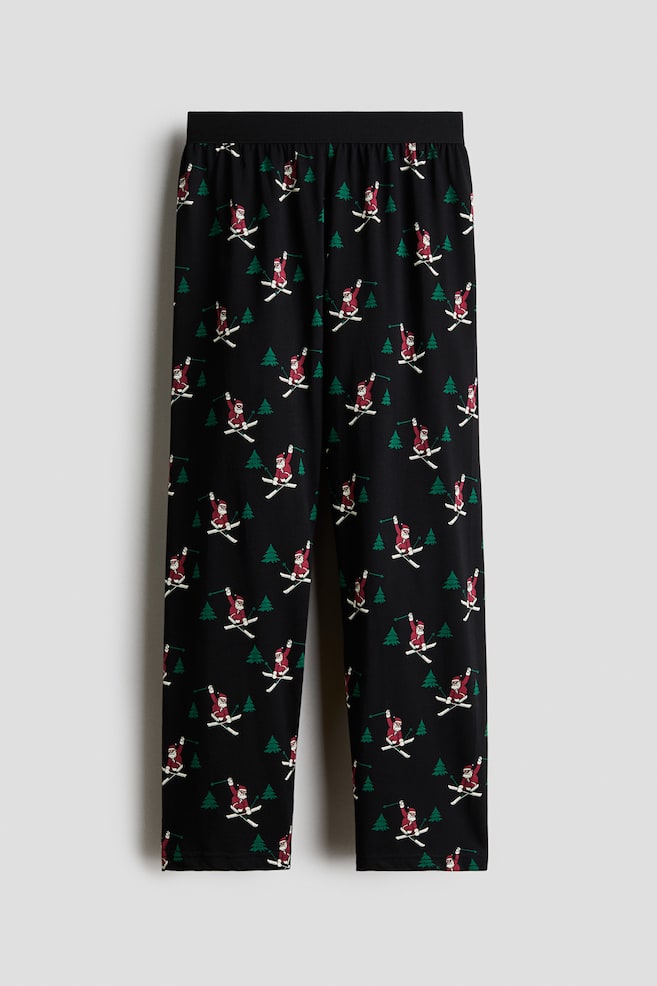 Pyjamasbukser i svart bomullsjersey vises forfra, med en bred, svart elastisk linning og rette ben. Stoffet har et heldekkende mÃ¸nster av Skiing Santa Claus-figurer og smÃ¥ grÃ¸nne furutrÃ¦r. - 1