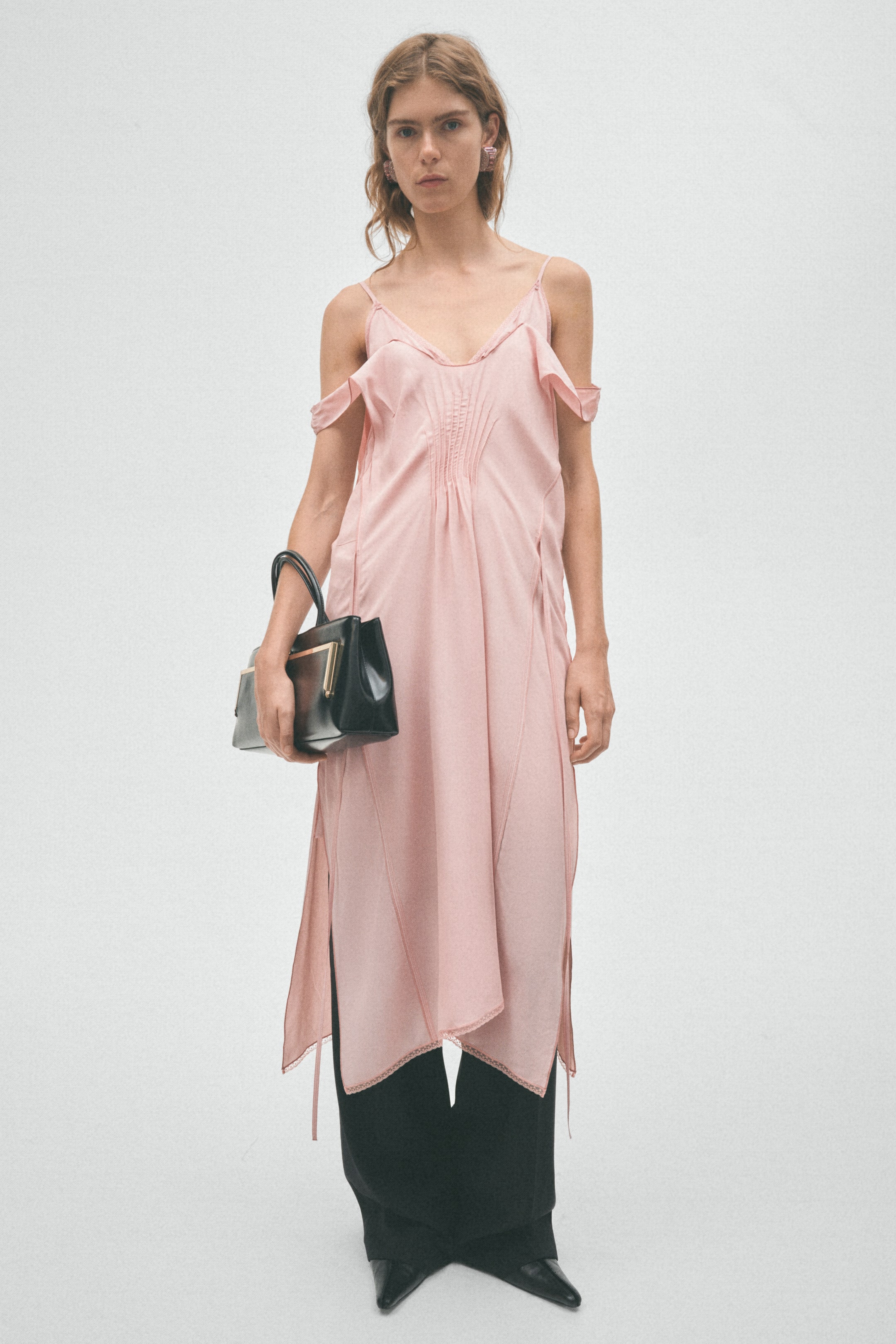 View larger image: The light pink slip dress, with spaghetti straps, off-shoulder ruffles, central pintucks, and an asymmetrical hem, is worn by the model. It is layered over black wide-leg pants, and the model is holding a black structured handbag.