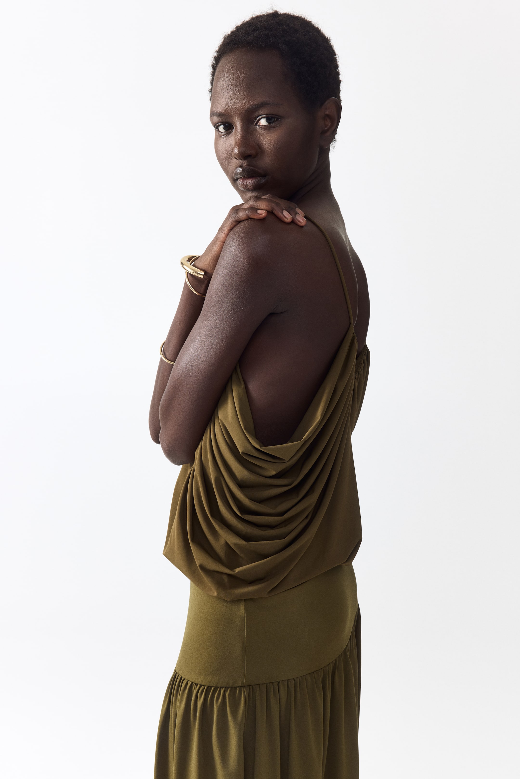 Grotere afbeelding bekijken: Een olijfgroene jurk met een camisole-achtige lijfje en een diep gedrapeerde cowl rug, die volumineuze stofplooien creëert, en een lange rok met een geplooide rucheszoom, getoond vanuit een zij-achteraanzicht.