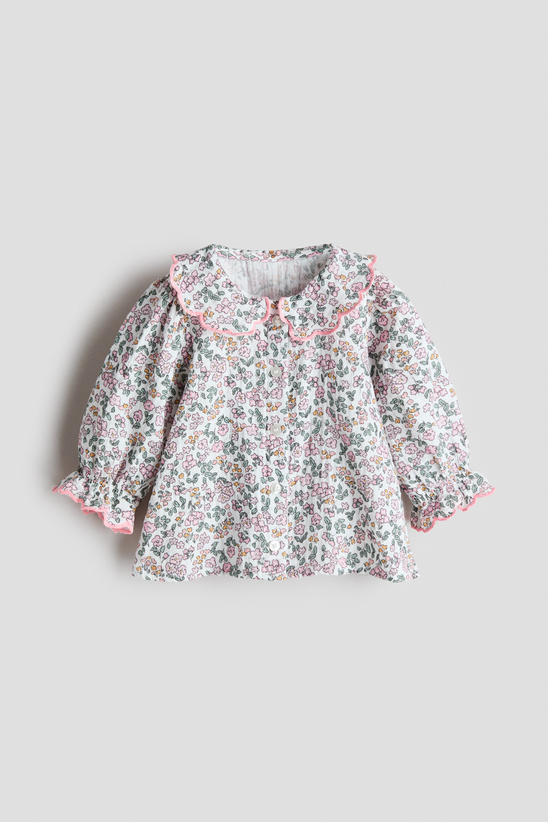 View larger image: A floral blouse is presenting FRONT, featuring a white base with a small pink, green, and yellow floral pattern. It is designed with a scalloped Peter Pan collar and ruffled cuffs, both accented with pink trim, and is closing down the front with white buttons.