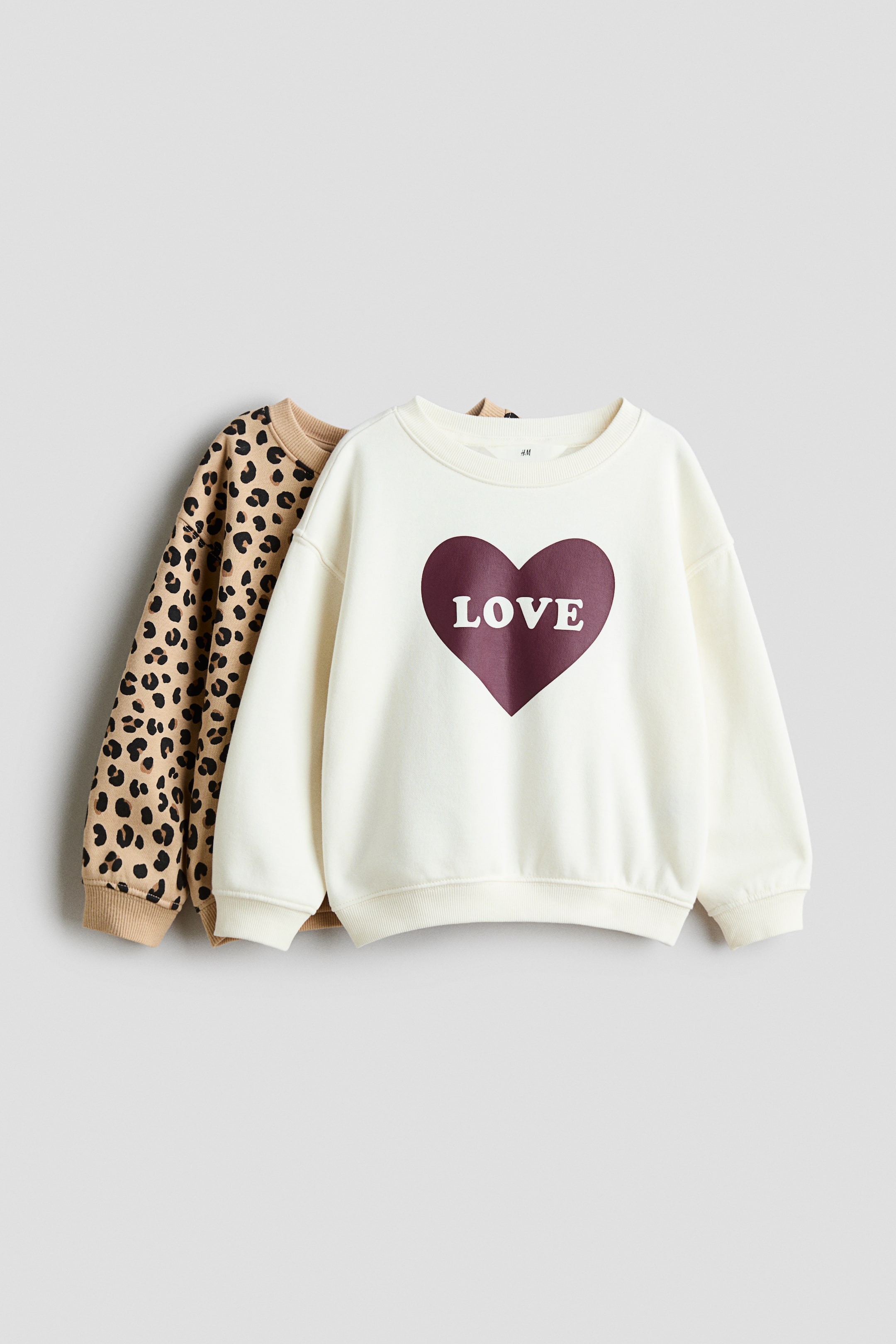 View larger image: Two long-sleeved sweatshirts are layered together, with the front off-white crewneck sweatshirt featuring a large burgundy heart graphic and the word "LOVE" printed in white. The second sweatshirt, layered behind, features an all-over leopard print pattern with solid tan ribbed trims.