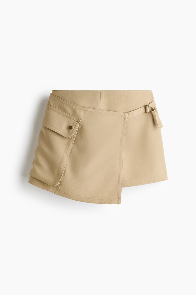 A beige wrap mini skirt is facing front, showcasing an asymmetrical front panel and a large cargo pocket with a snap-button flap on the right side. The skirt is featuring a flat waistband and an adjustable side strap with a metal buckle on the left. - 1