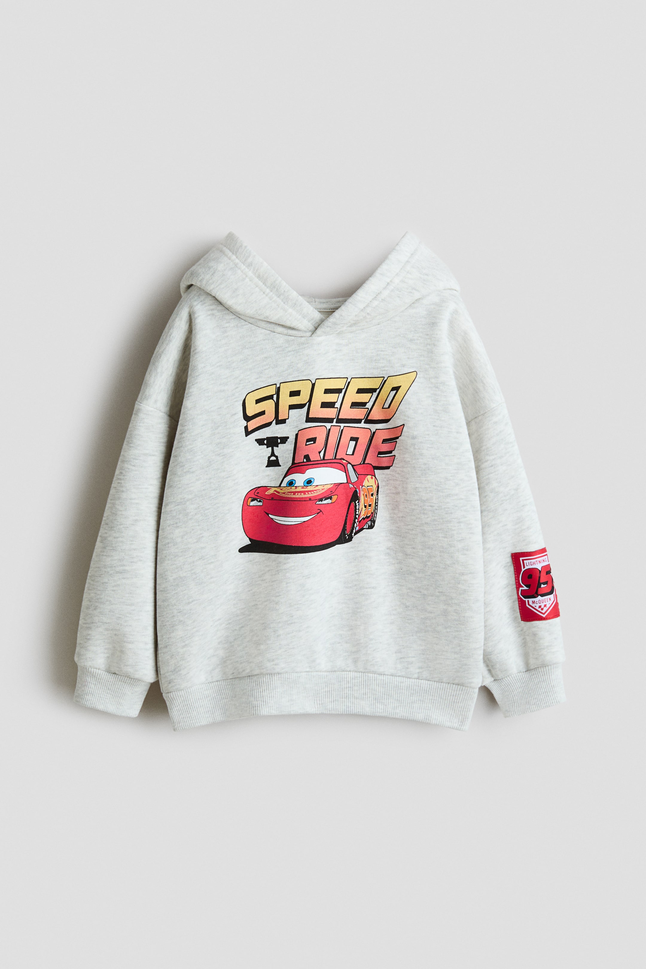Ver imagem maior: Um hoodie estampado de corte largo apresenta a frente em cinzento claro mesclado, com uma grande estampa gráfica do Lightning McQueen com o texto "SPEED RIDE". O hoodie apresenta também punhos canelados, uma bainha canelada e um patch vermelho "95 Lightning McQueen" na manga esquerda.