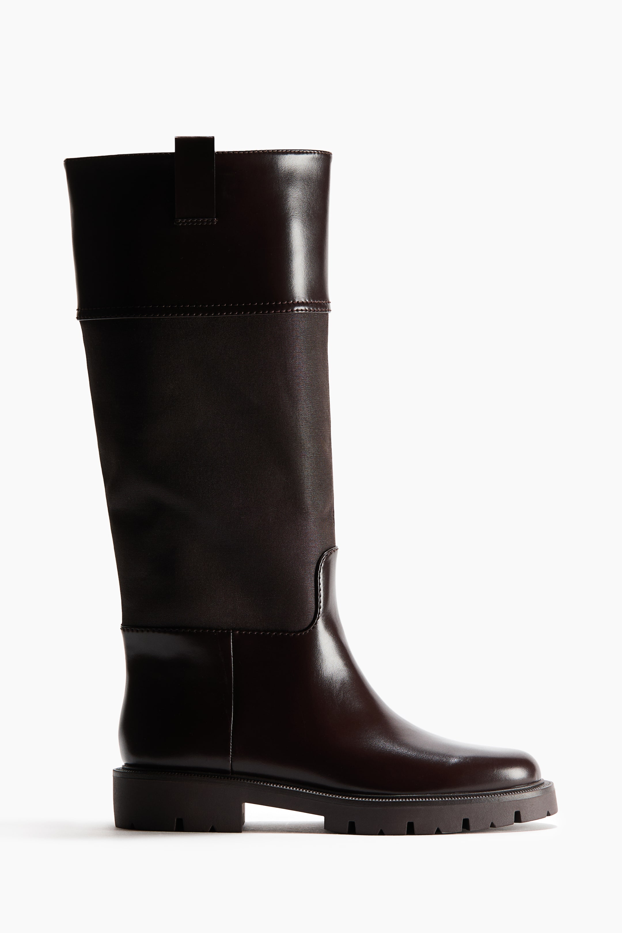 View larger image: A dark brown knee-high boot is shown in side profile, featuring a glossy leather foot and upper cuff with a small pull tab at the top. The mid-shaft is made from a contrasting matte dark brown material and finished with a thick lug sole.