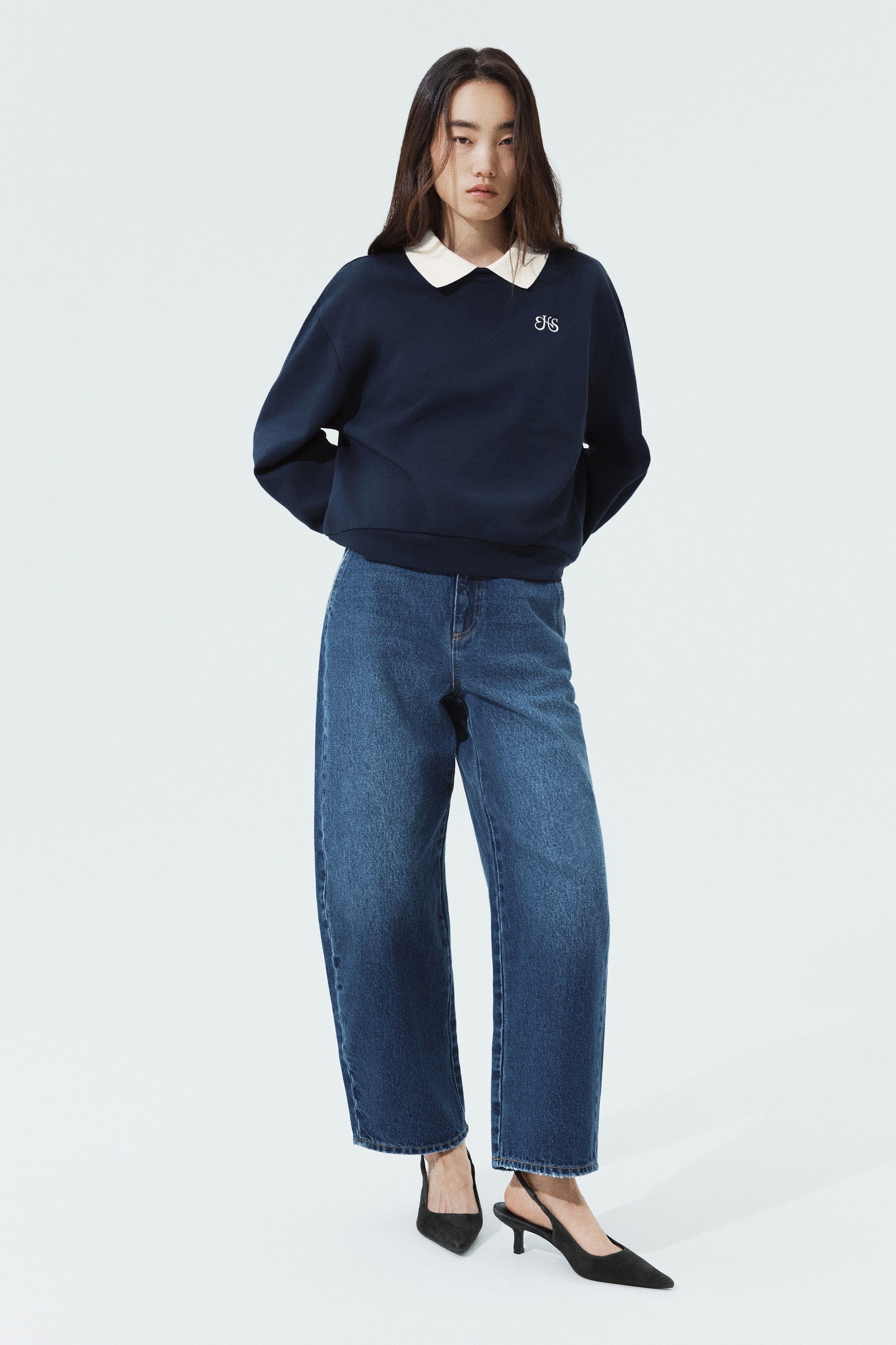 이미지 확대 보기: 모델이 대비되는 흰색 칼라, 드롭 숄더, 가슴 부분의 작은 흰색 자수 디테일이 돋보이는 네이비 블루 스웨터를 착용하고 있습니다. 긴 소매 스웨터는 와이드 레그 블루 데님 진과 블랙 슬링백 힐과 함께 스타일링되었습니다.