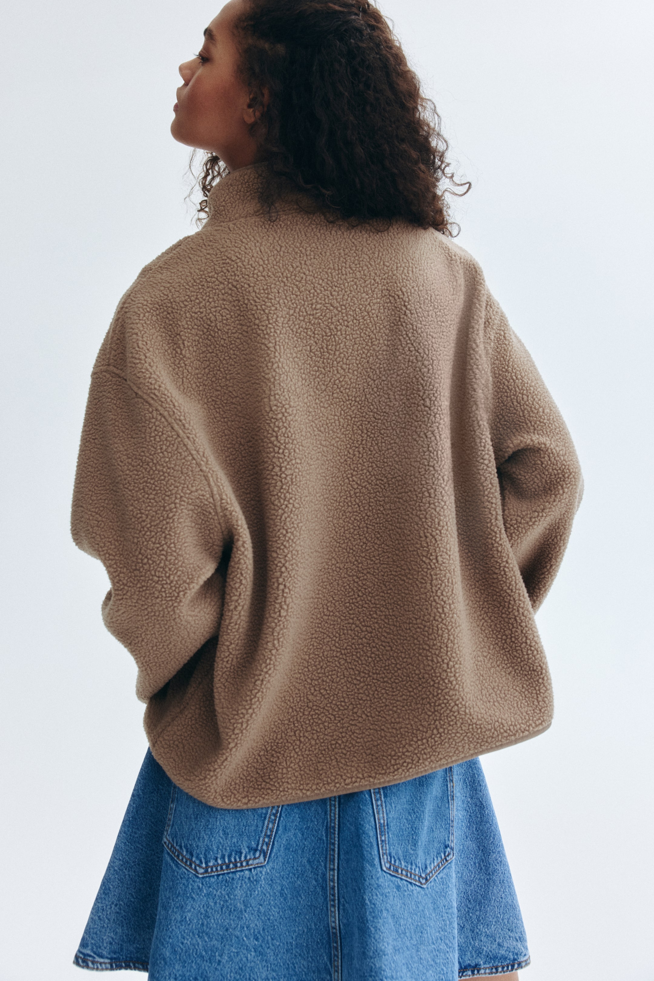 Agrandir l'image: Un haut en polaire teddy marron est porté par le mannequin, vu de dos. Il présente un col montant, des manches longues et une silhouette décontractée, avec son matériau texturé visible au-dessus d'une jupe en denim bleu.