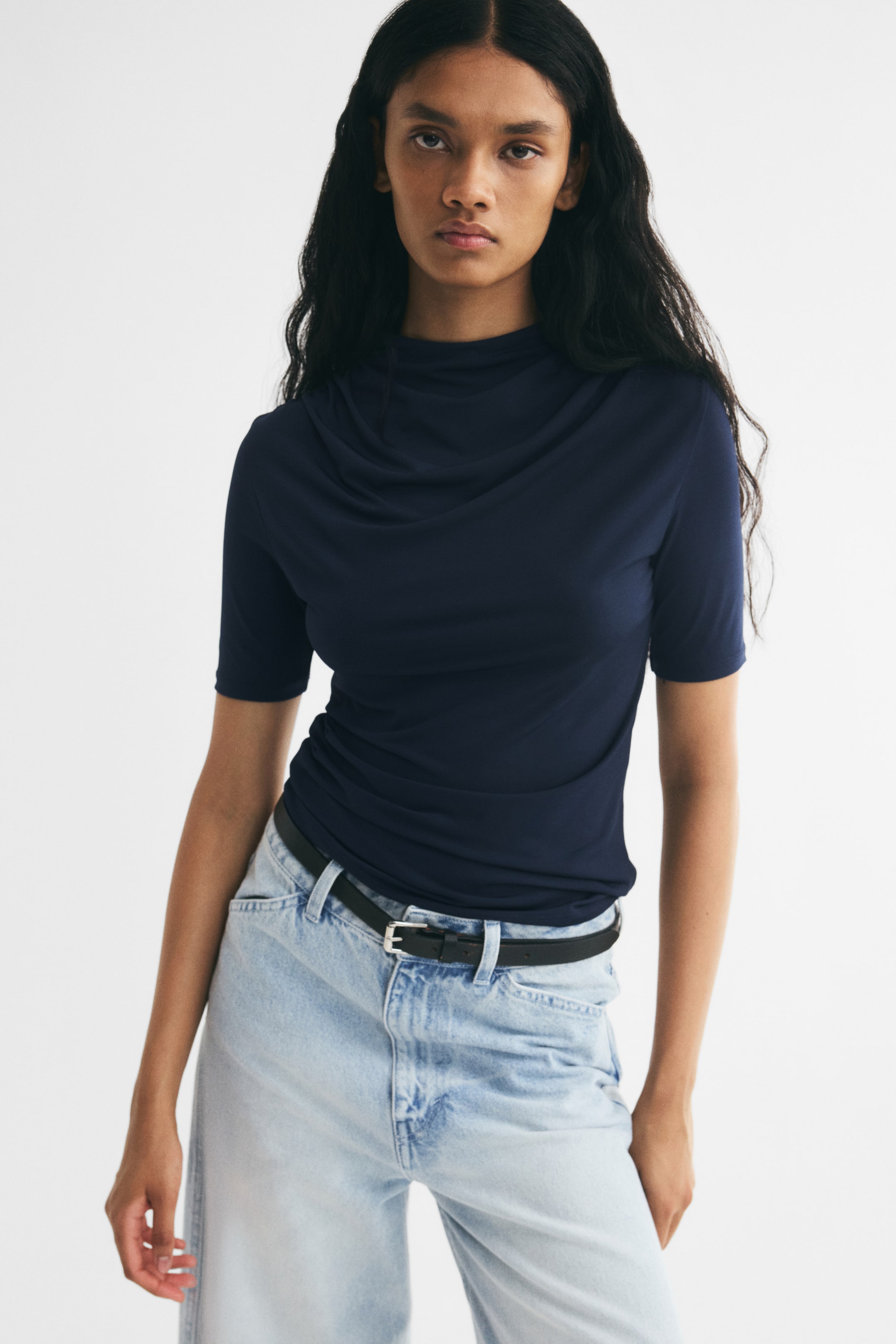 Ampliar la imagen: La modelo lleva una blusa ajustada en azul marino oscuro de punto elástico, con cuello alto drapeado y frunces en los hombros, y mangas cortas. La prenda se combina con jeans azules de lavado claro y un cinturón negro delgado.