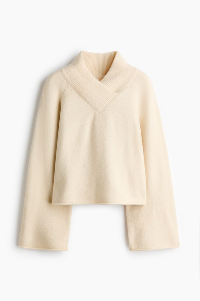 A cream-coloured knit jumper is shown from the front, featuring a wide, ribbed crossover V-neck collar and long, flared sleeves. The jumper has a relaxed, slightly cropped silhouette with a clean, straight hem. - 1