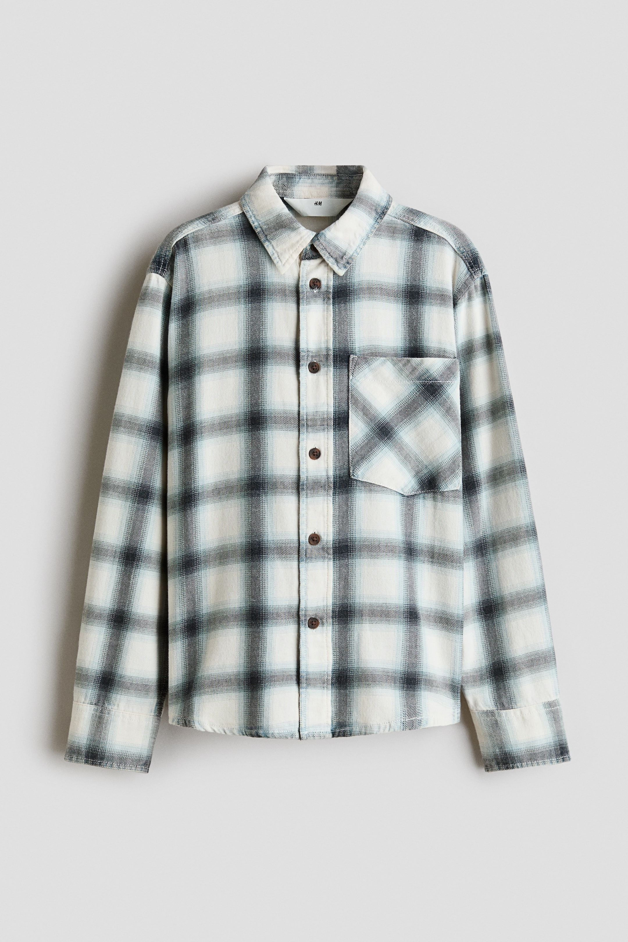 Ver imagen más grande: Una camisa de cuadros de manga larga cuelga orientada hacia el frente, mostrando un estampado a cuadros en tonos de blanco, gris y azul claro. La camisa presenta un clásico cuello de punta, cierre frontal con botones de color marrón oscuro y un único bolsillo de parche en el pecho izquierdo.