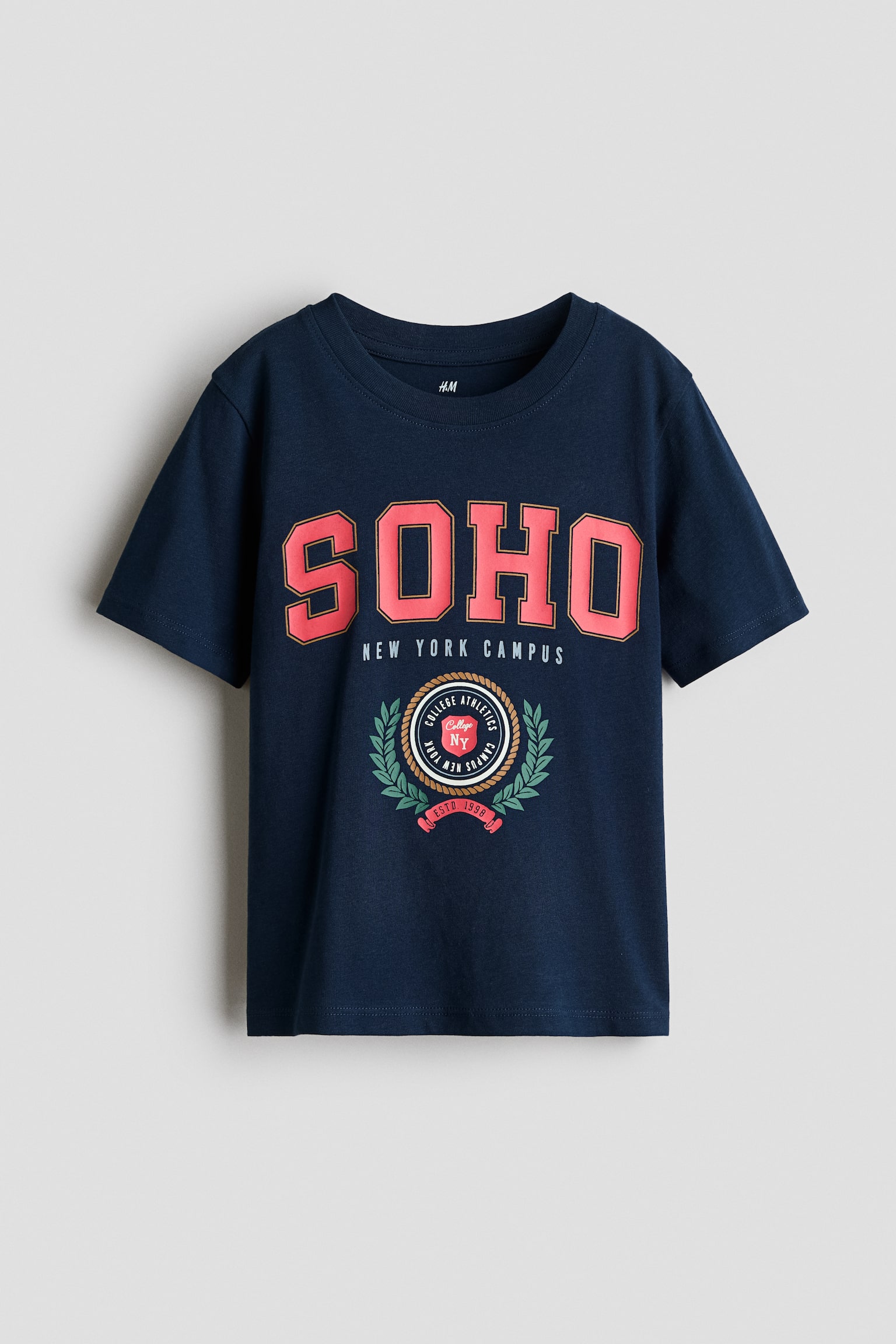 A navy blue cotton jersey t-shirt is shown front-facing, featuring a round ribbed neckline and short sleeves with softly dropped shoulders. The chest displays a printed collegiate graphic with "SOHO" in large pink block letters above the text "NEW YORK CAMPUS" and a circular athletic crest.