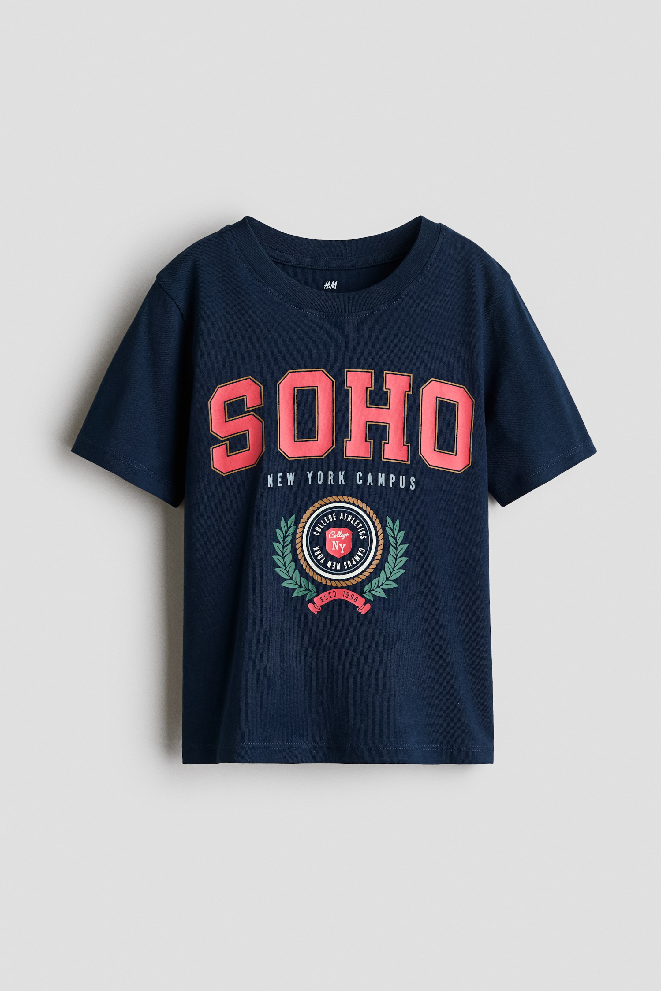 View larger image: A navy blue cotton jersey t-shirt is shown front-facing, featuring a round ribbed neckline and short sleeves with softly dropped shoulders. The chest displays a printed collegiate graphic with "SOHO" in large pink block letters above the text "NEW YORK CAMPUS" and a circular athletic crest.